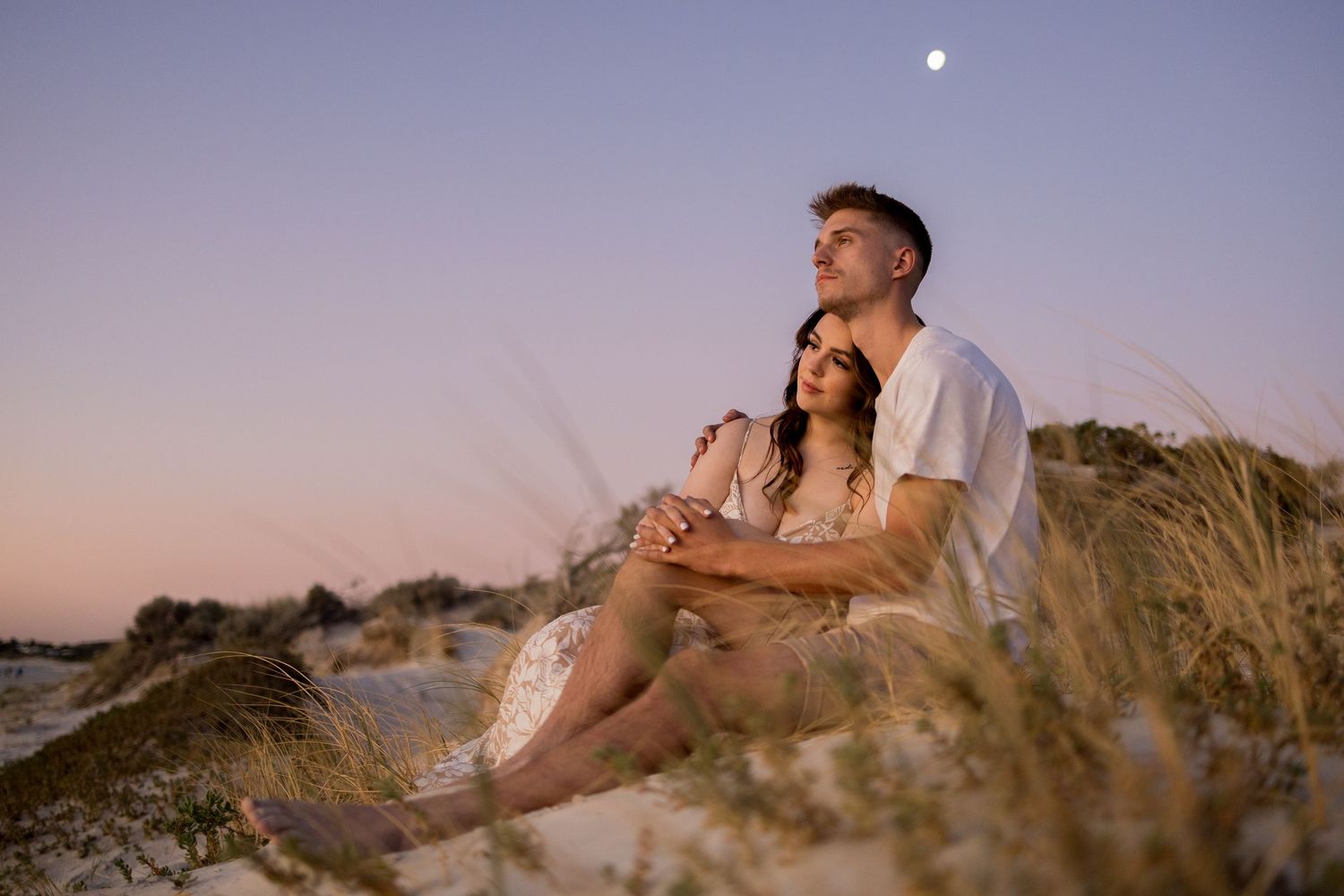 Young couple sitting on sand dunes watching moon rise at dusk.