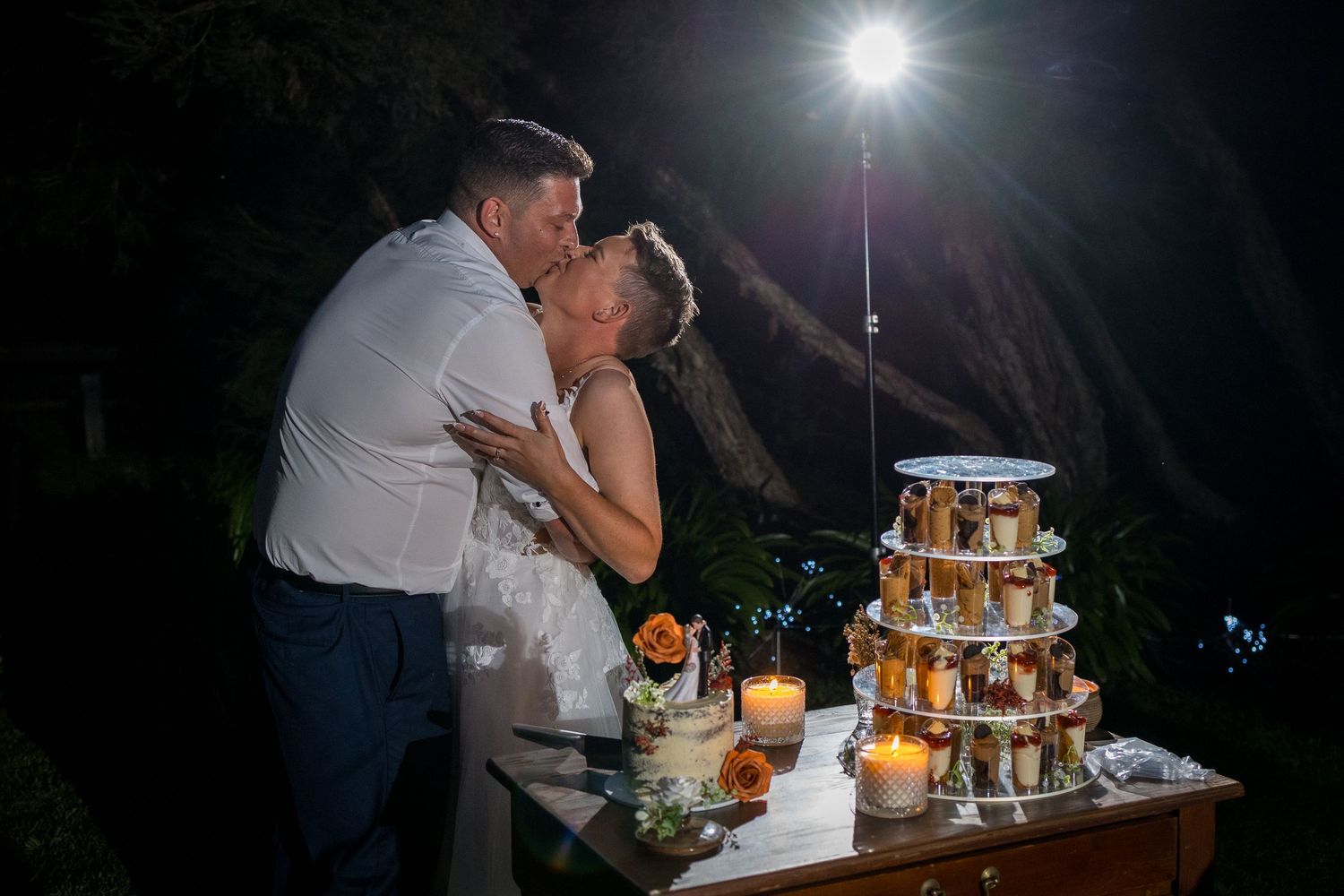 Bride and groom share a romantic kiss beside a tiered wedding cake display at an outdoor night reception.