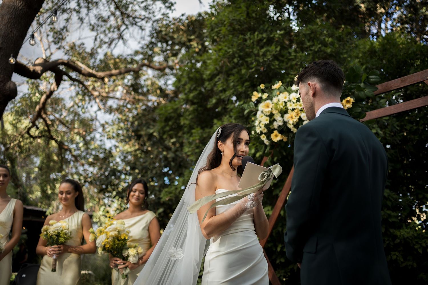 Bride reading vows to groom at outdoor wedding ceremony with bridesmaids and yellow floral arch in background.