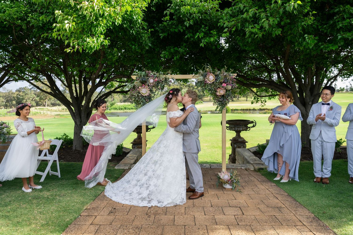 Bride and groom share first kiss at outdoor wedding ceremony under trees with bridal party beside them.