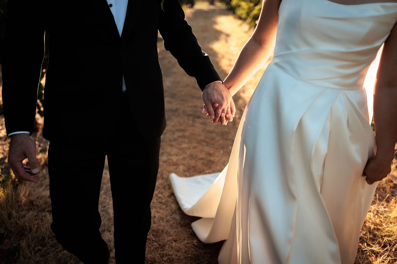 Close-up of bride and groom holding hands in warm sunset light, wedding attire detail shot.