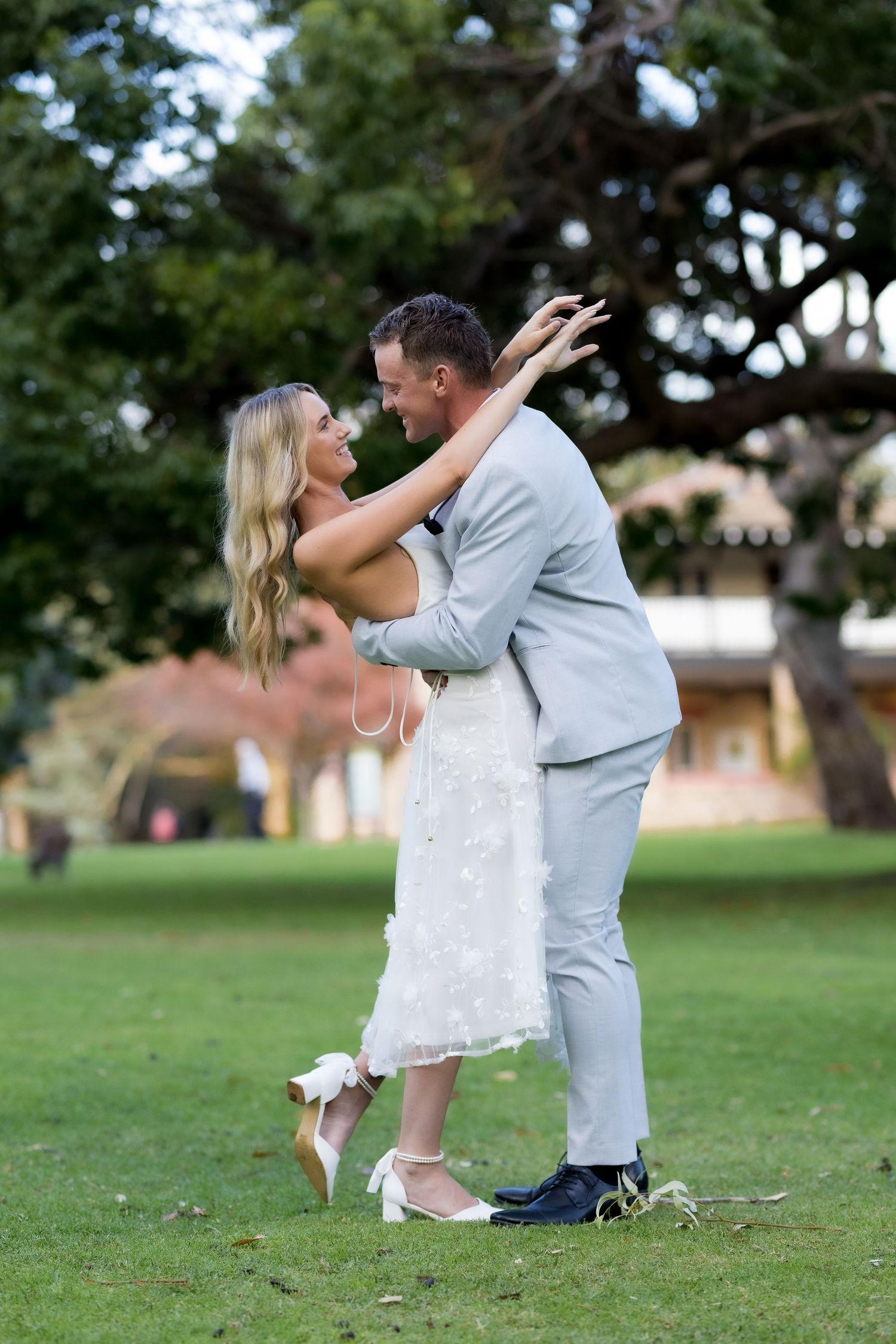 Bride and groom dancing and laughing together in sunny park setting.