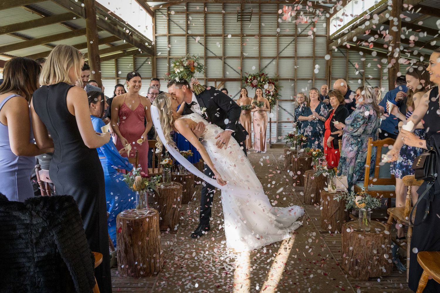 Groom dips bride for a kiss as confetti fills the air during a rustic barn wedding ceremony with guests. at Quarry Farm