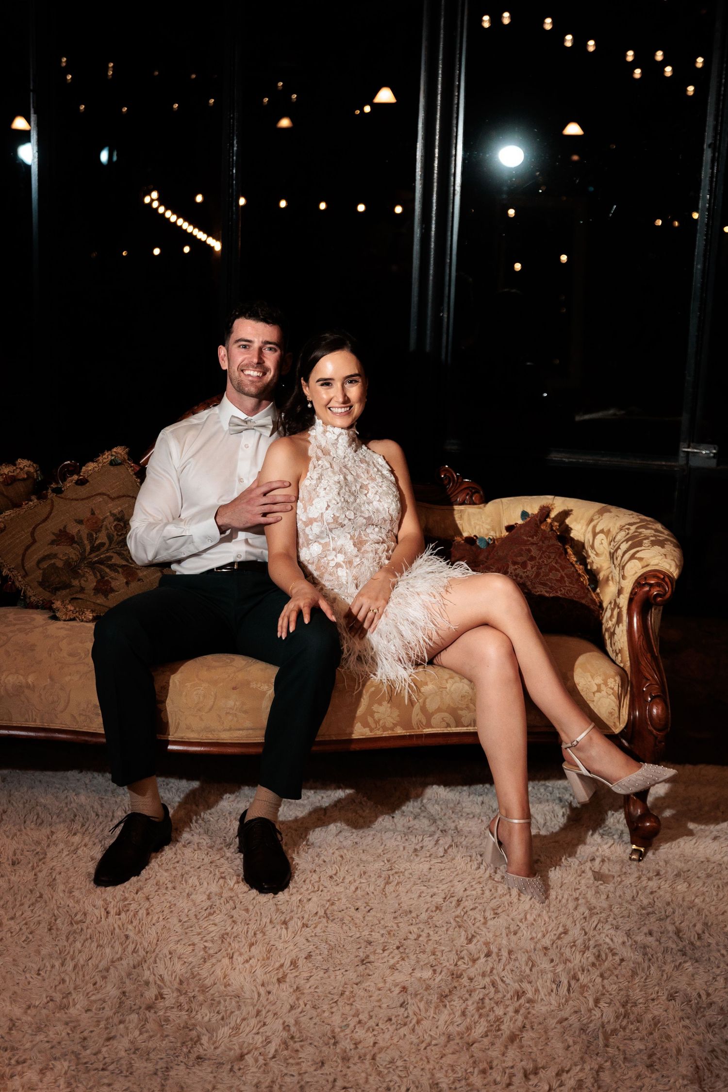 Elegant couple sitting on vintage sofa in formal attire, woman in white feather dress, man in bow tie, at night event.