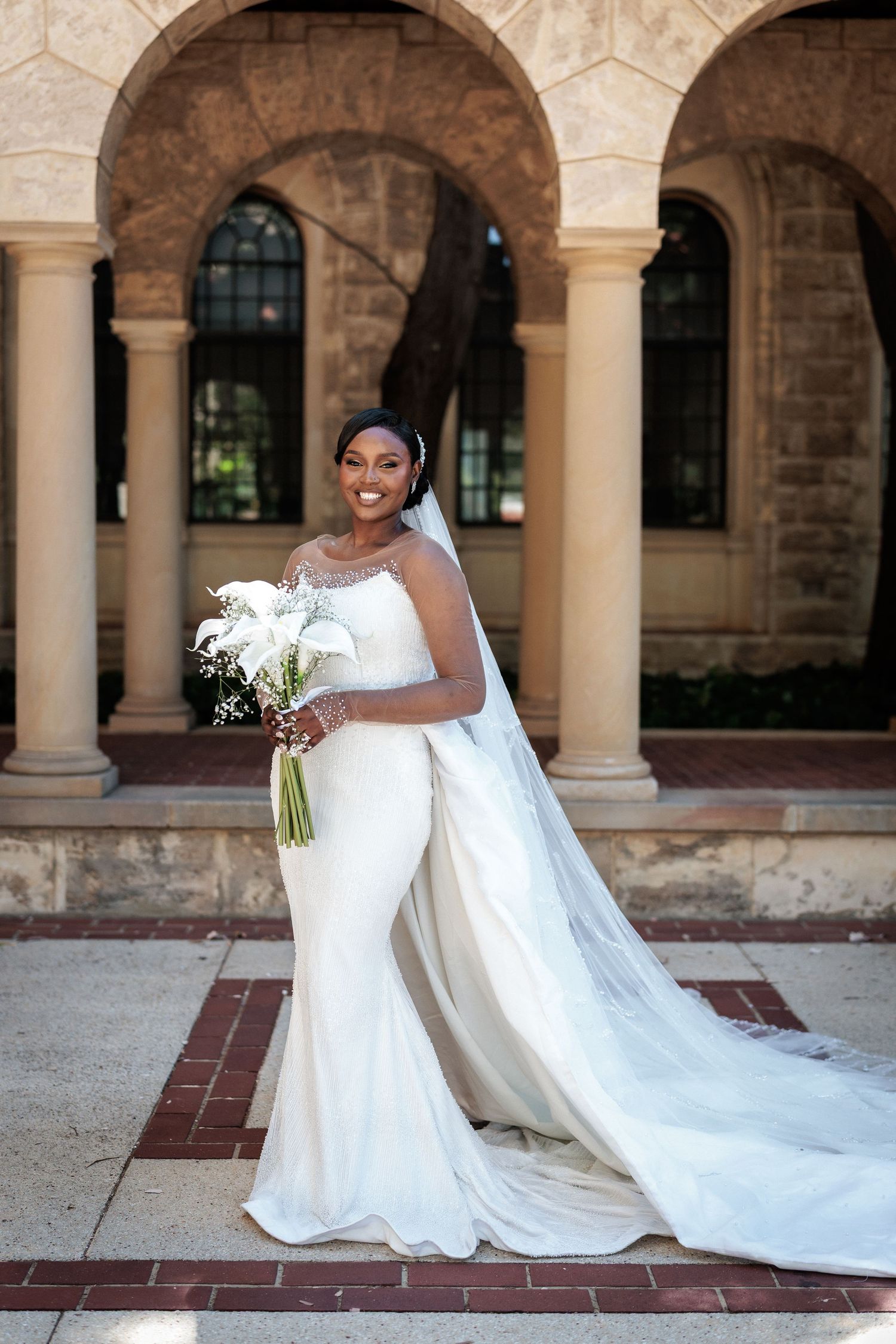 Radiant bride in fitted white gown with long veil holds bouquet under stone archway columns.