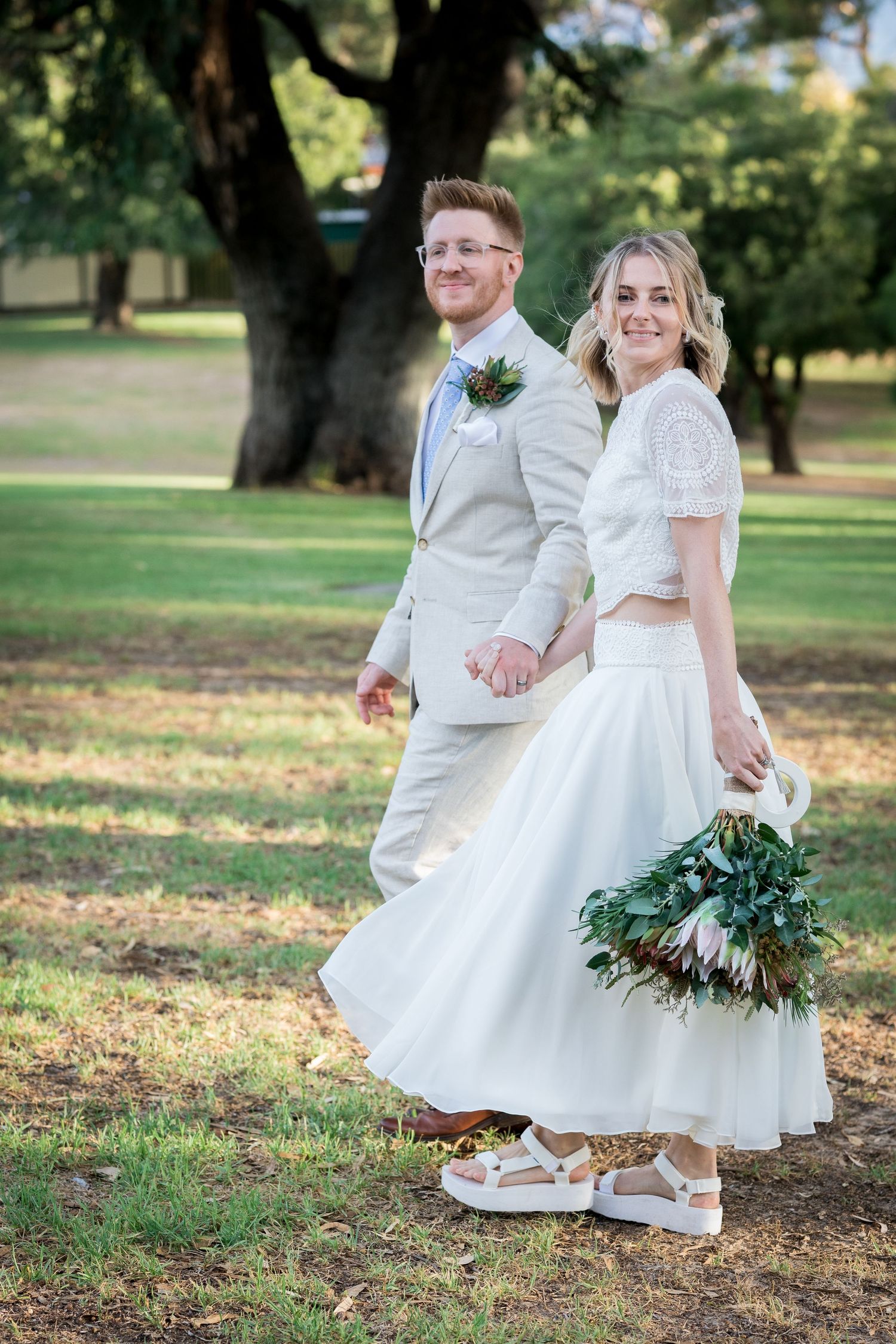 Smiling bride and groom in casual chic attire hold hands while strolling through a sunlit park.