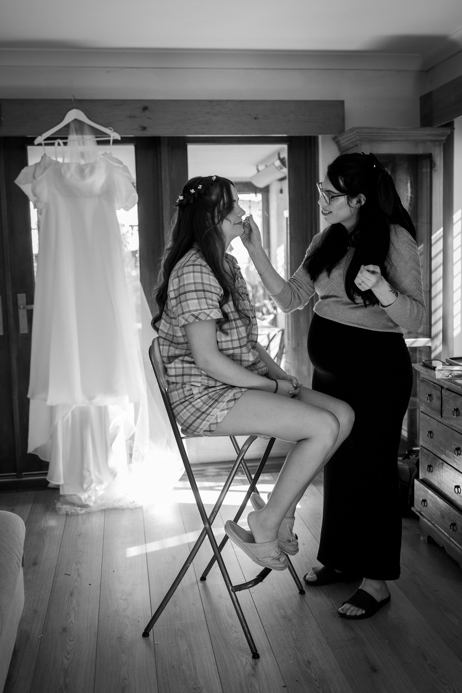 Black and white photo of bride getting makeup done by bridesmaid, wedding dress hanging in background.