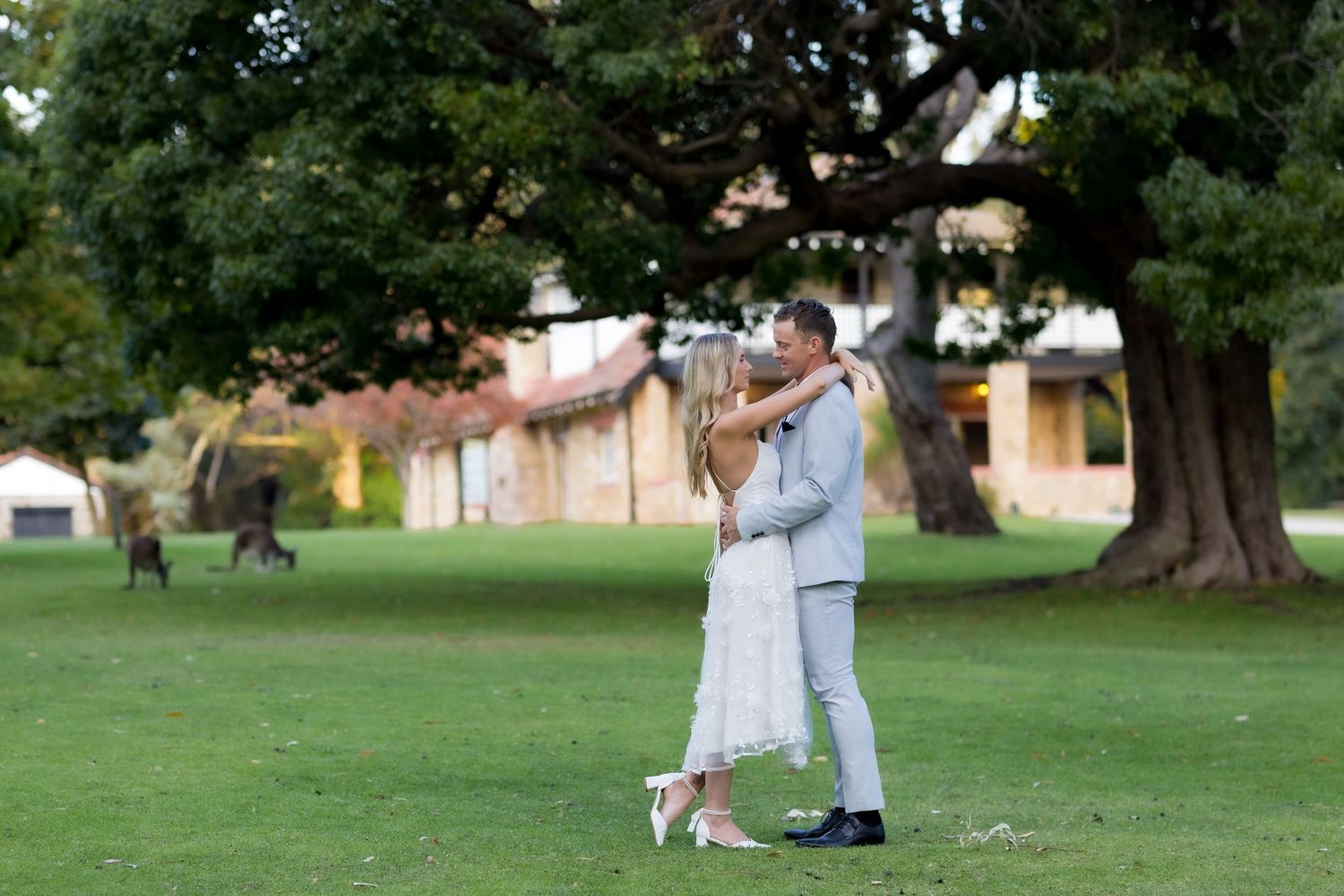 Couple embracing and dancing together on lush green lawn beside large tree at outdoor wedding venue.