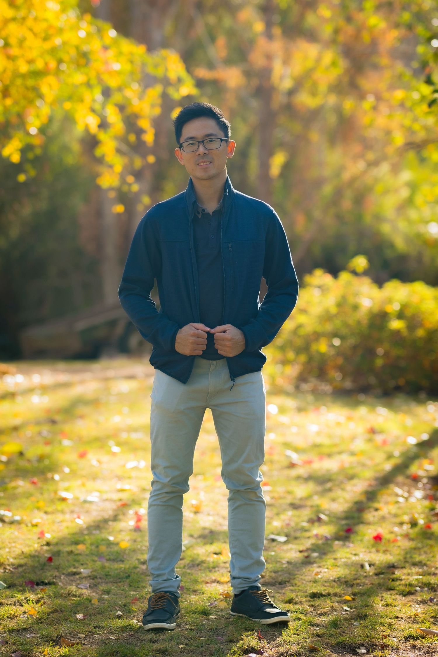 Young man in blue jacket and glasses stands confidently in a sunlit autumn park with colorful foliage.