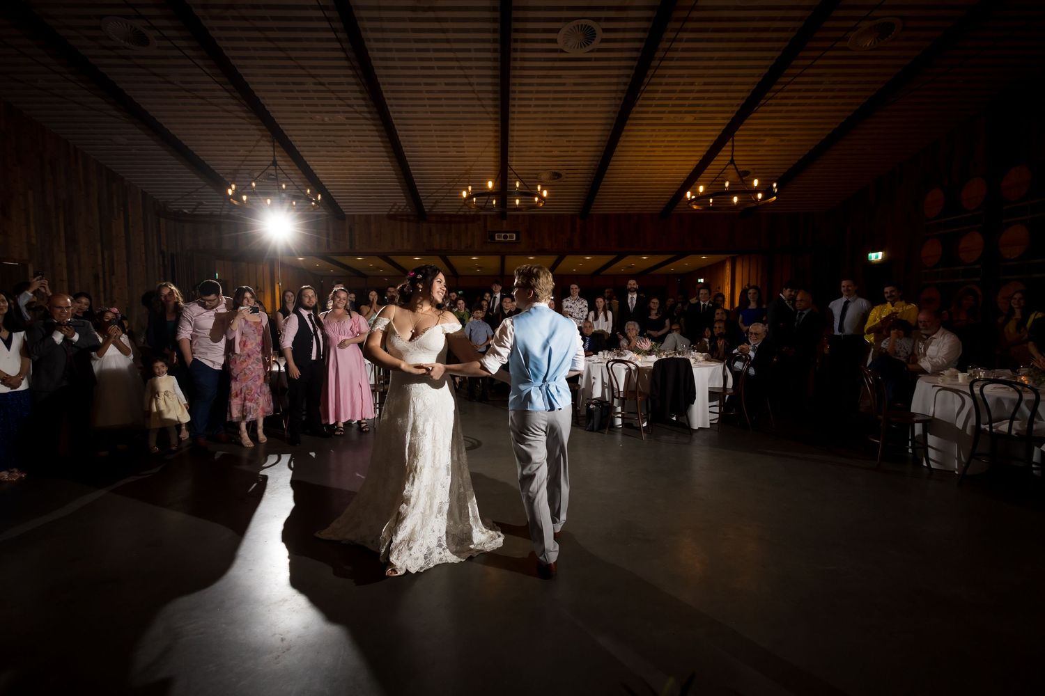 Bride and groom share first dance on dimly lit reception dance floor surrounded by celebrating guests.