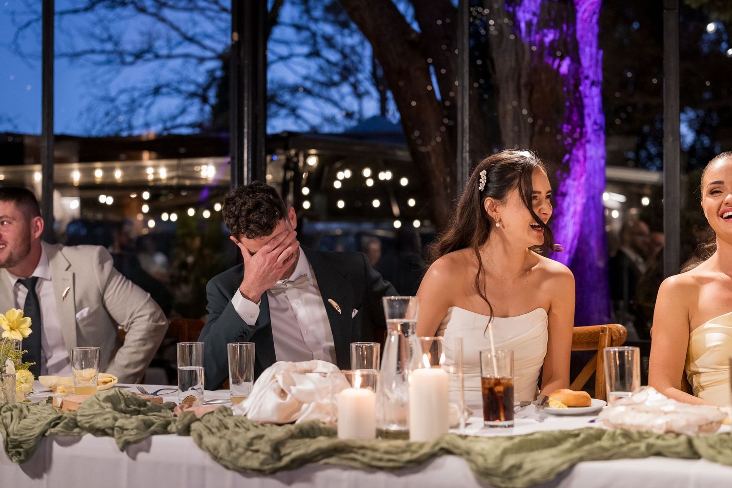 Bride and groom laugh at head table during evening wedding reception with candles and string lights.