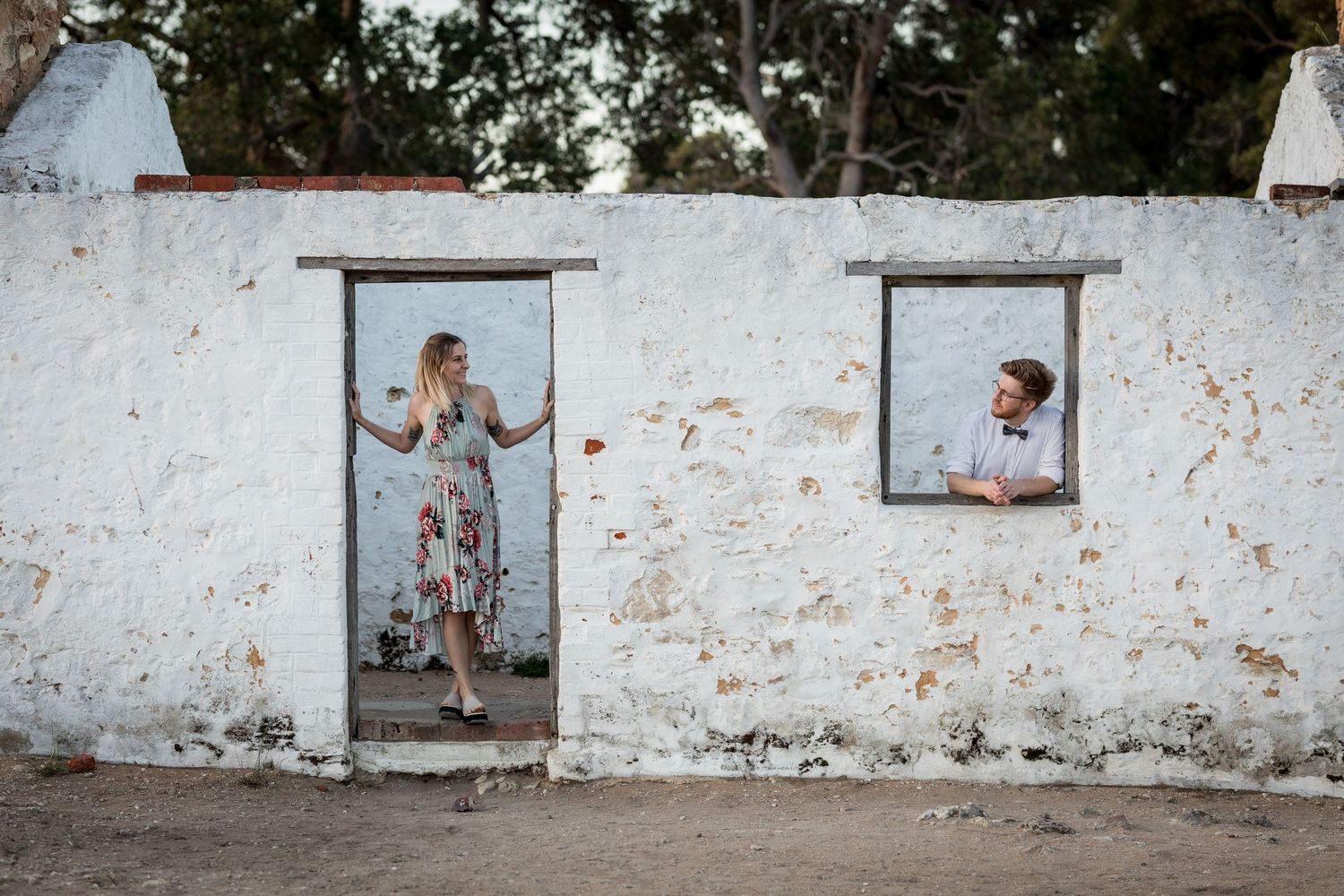 Two people pose playfully in doorways and windows of a weathered white wall.