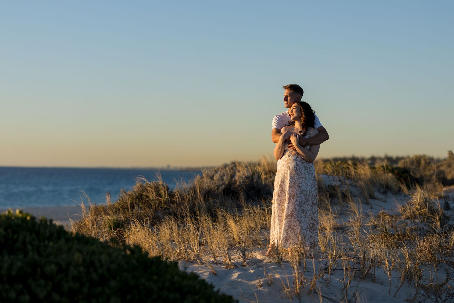 Couple embracing on sandy beach dunes at golden sunset with ocean view in background.