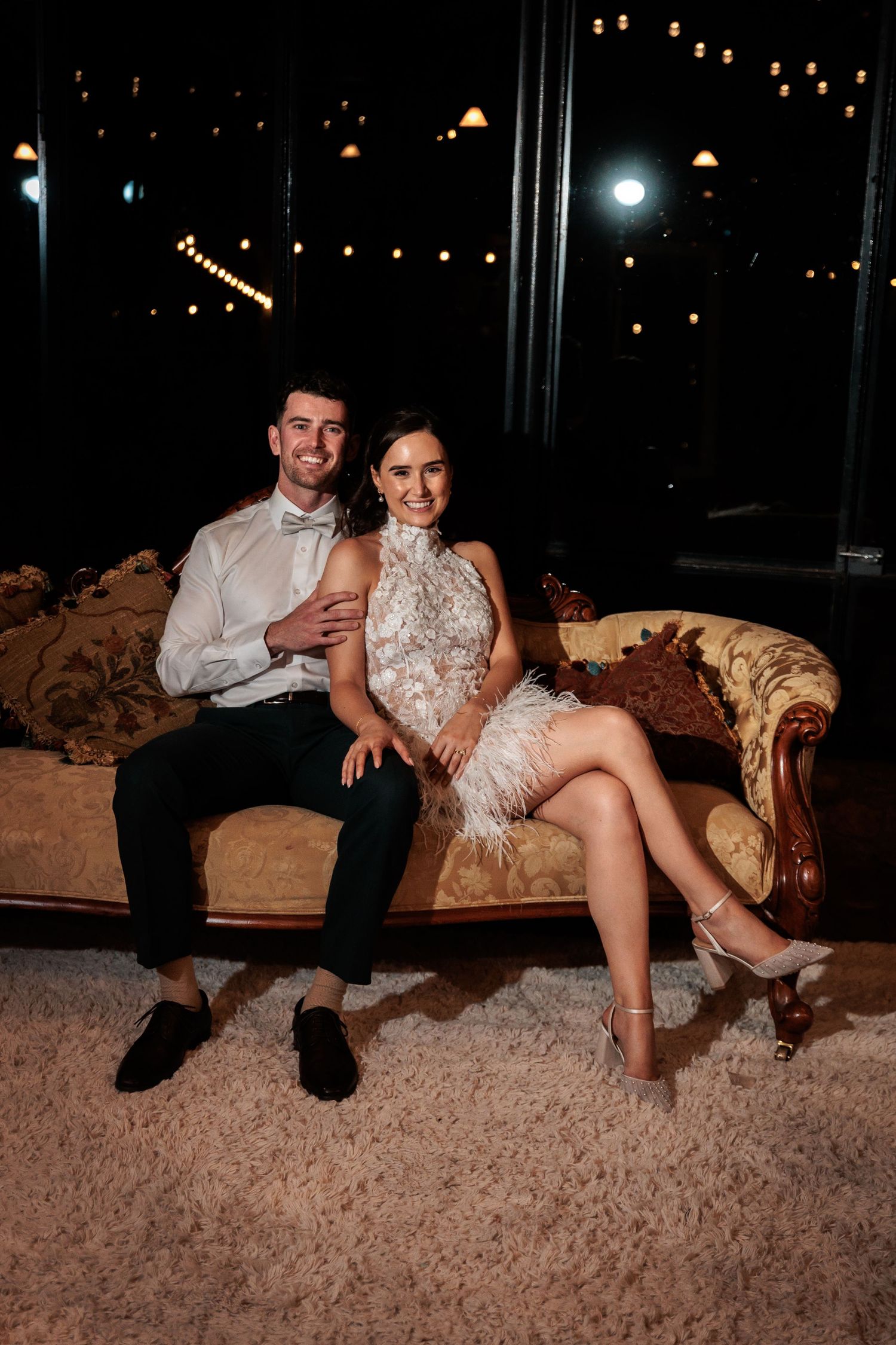 Elegant couple sitting on ornate vintage sofa, man in white shirt and bow tie, woman in feathered dress, at night event.
