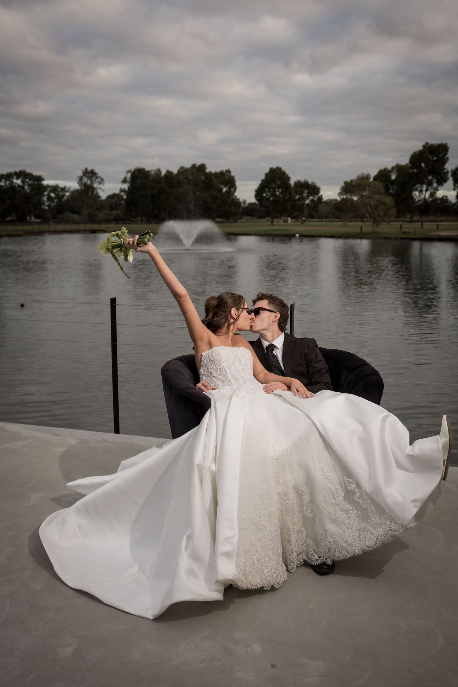 Bride and groom kissing on dock by lake, bride raising bouquet, voluminous white ball gown flowing.