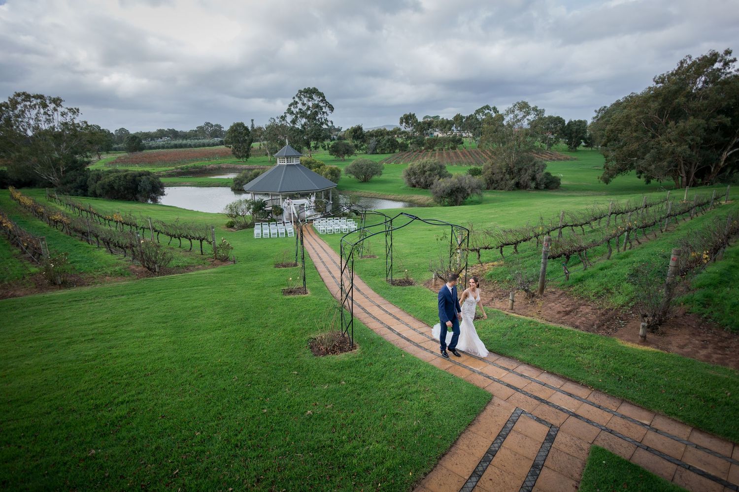 Sittella Winery-Aerial view of a wedding couple walking along a brick path through lush green vineyard with a gazebo and pond in background.