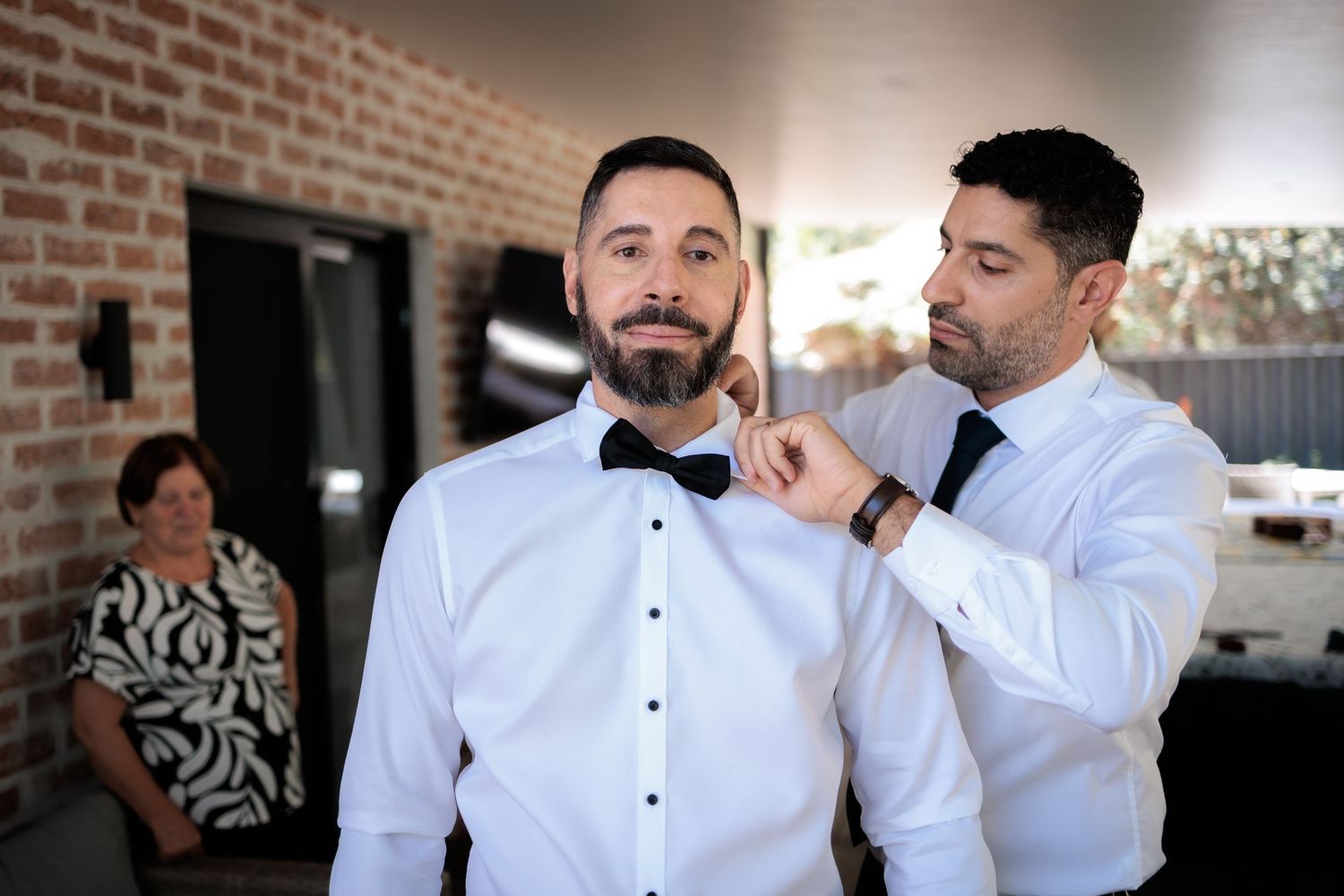 Groom getting bow tie adjusted by groomsman in white dress shirts before wedding ceremony.