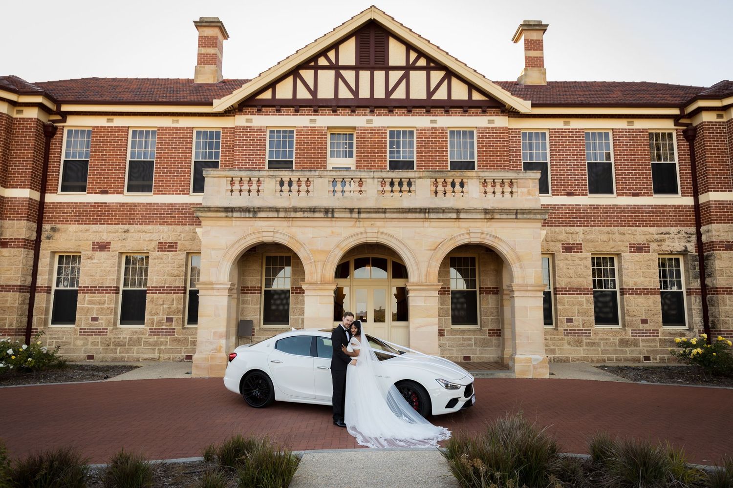 Bride and groom pose with white luxury car outside elegant red brick wedding venue at dusk at Montgomery Hall