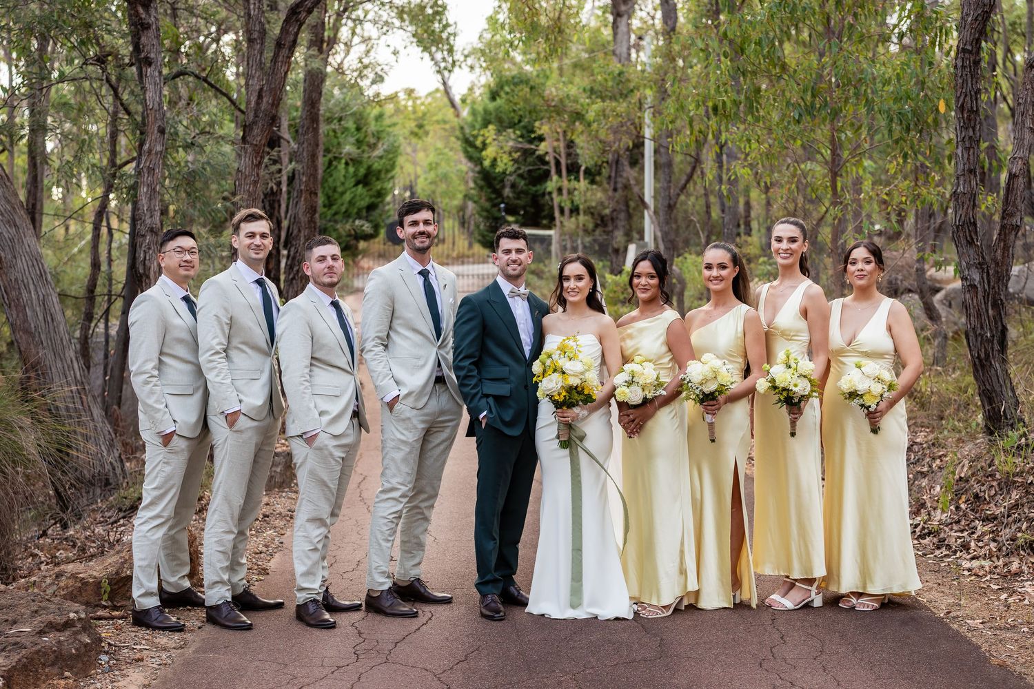 Wedding party posing on a forest path, groomsmen in tan suits, bridesmaids in yellow dresses holding white bouquets.