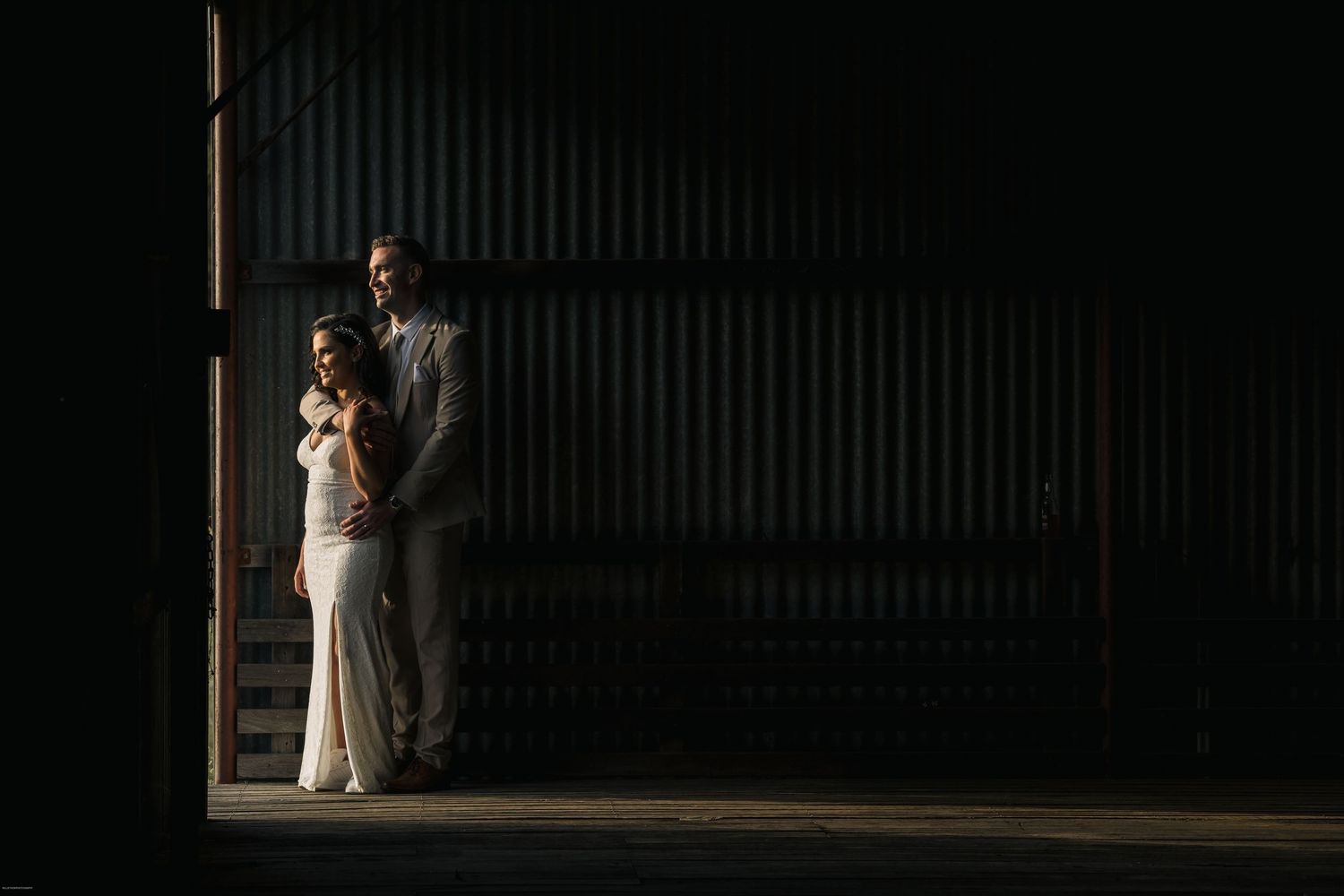 Bride and groom standing together in dramatic light inside a rustic barn during their wedding day.