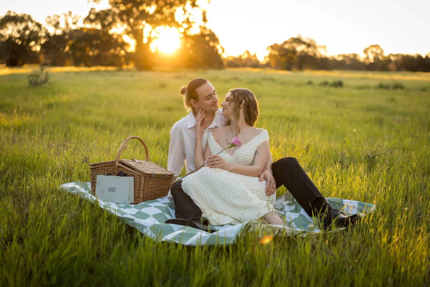 Couple sitting on a picnic blanket in a green field at golden sunset, sharing a romantic moment with a wicker basket nearby. at Guildford