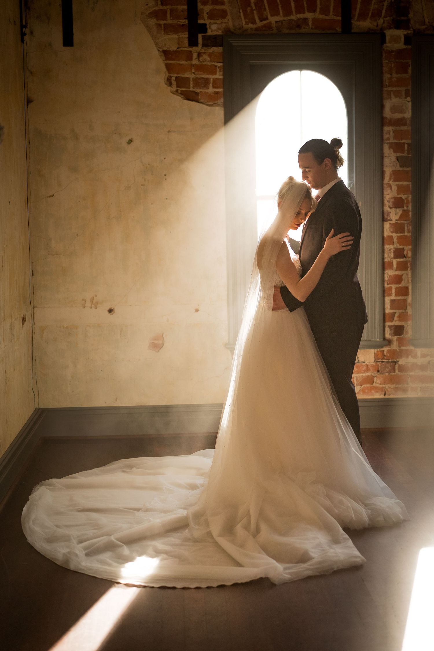Bride and groom embrace in soft sunlight streaming through arched window in rustic brick venue, wedding gown with long train.