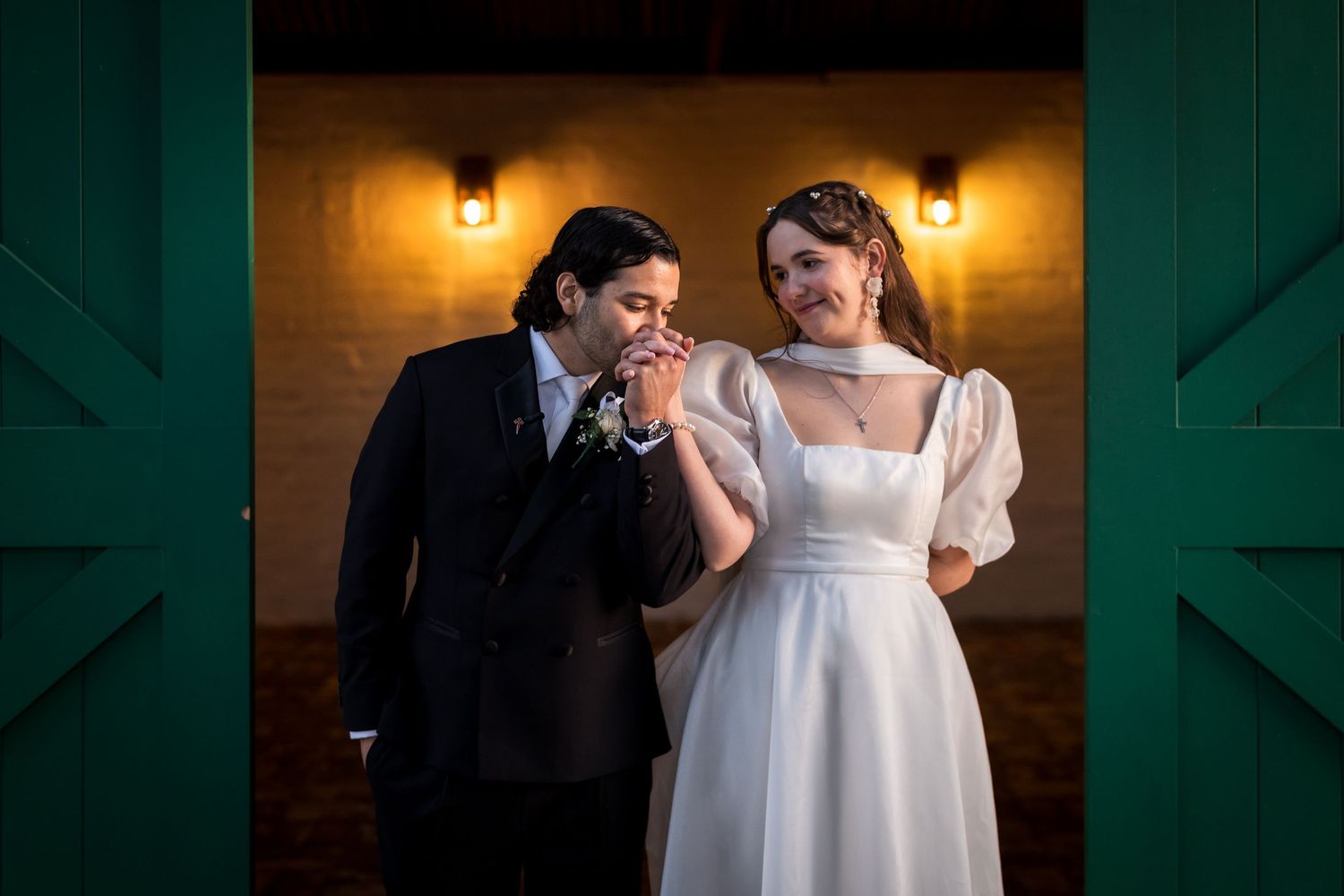 Groom kissing bride's hand in front of green barn doors at elegant wedding venue.