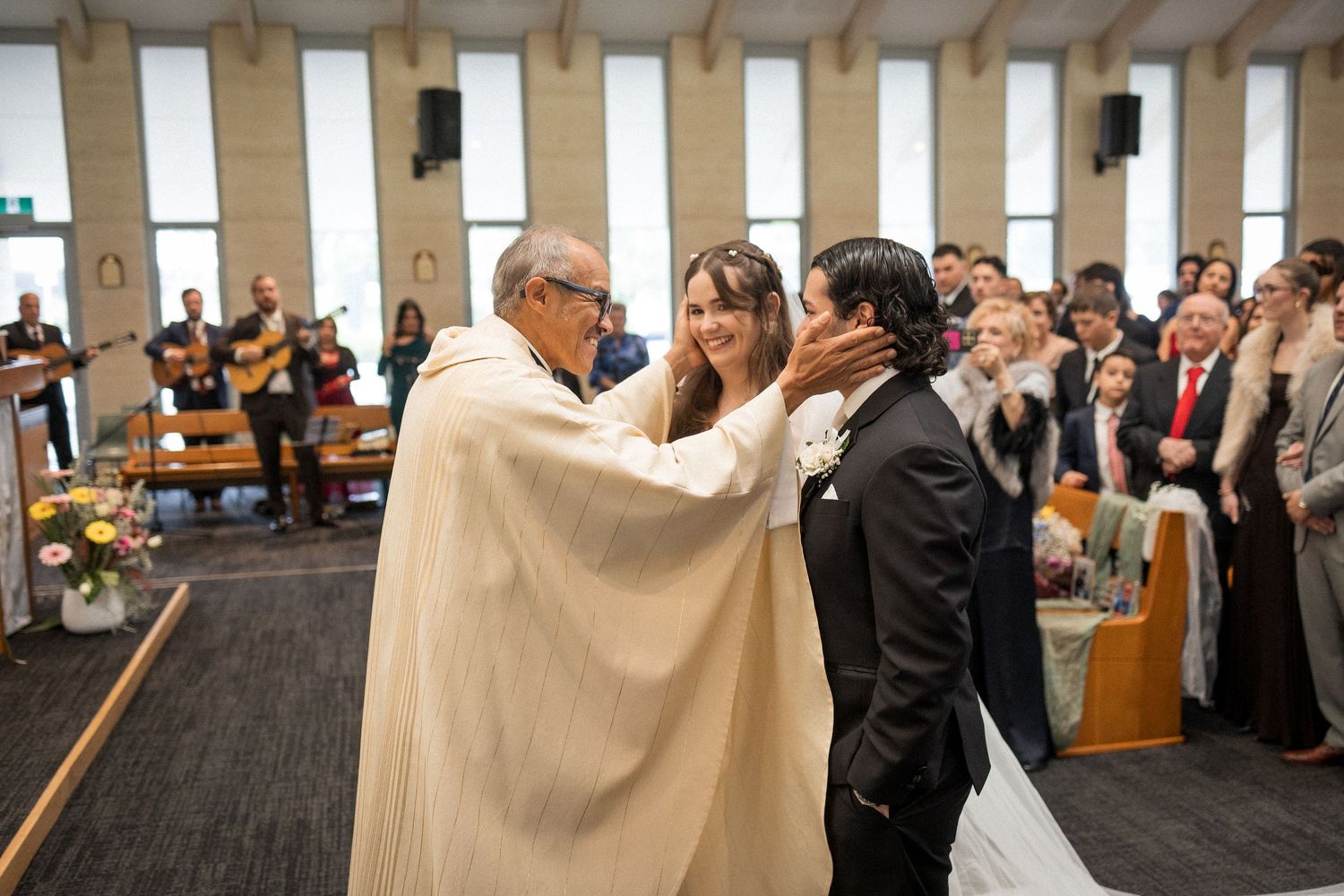 Priest blessing newlyweds during a wedding ceremony in a modern church with guests standing in the background.