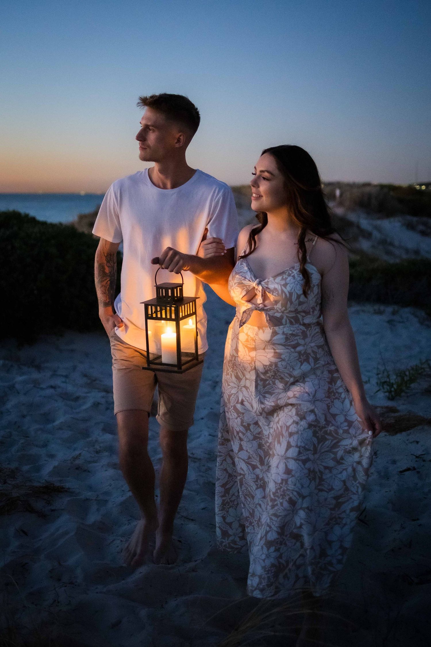Couple walking on beach at twilight holding glowing lantern together.