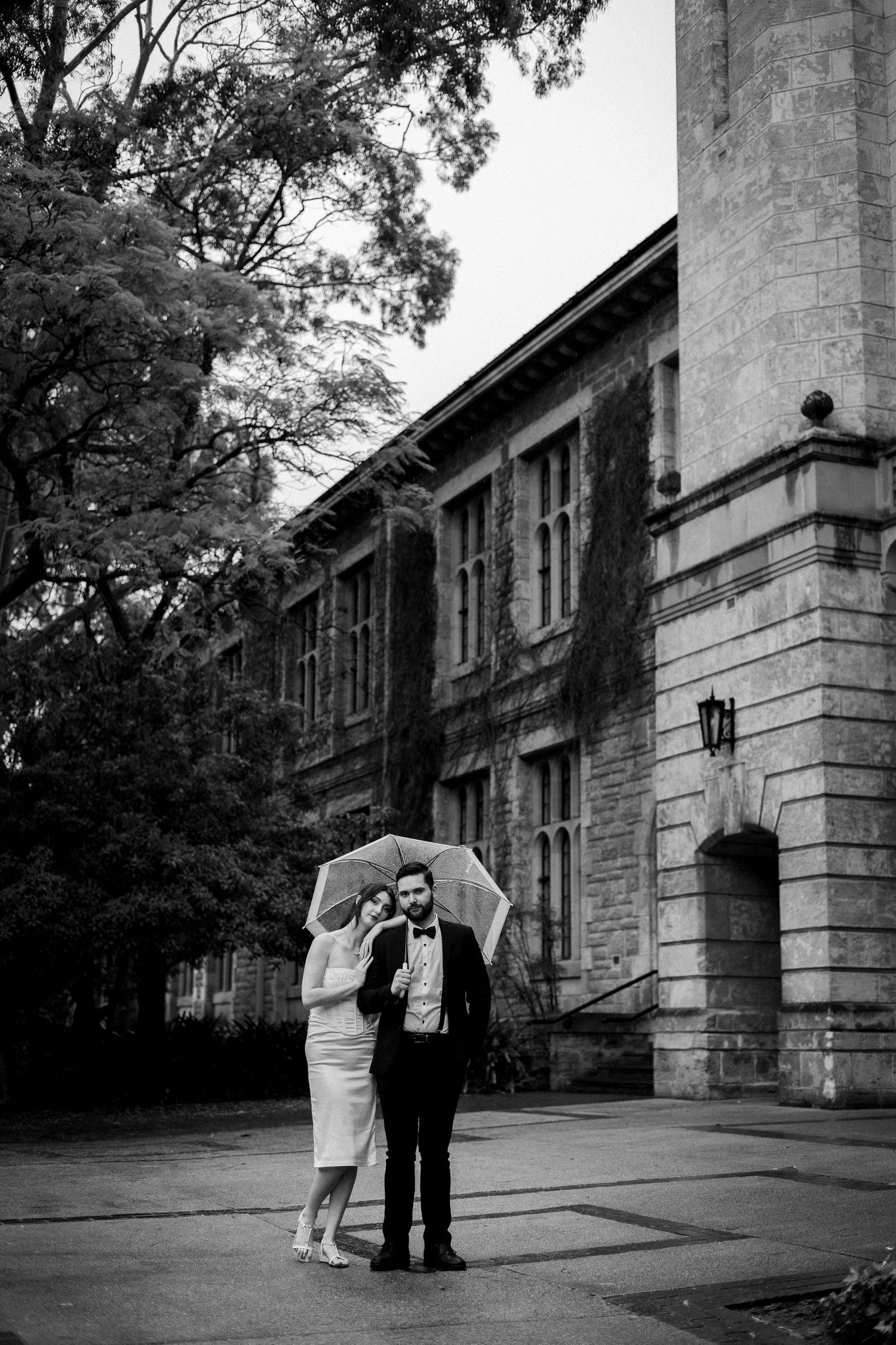 Black and white photo of a couple sharing an umbrella outside a historic stone building on their wedding day.