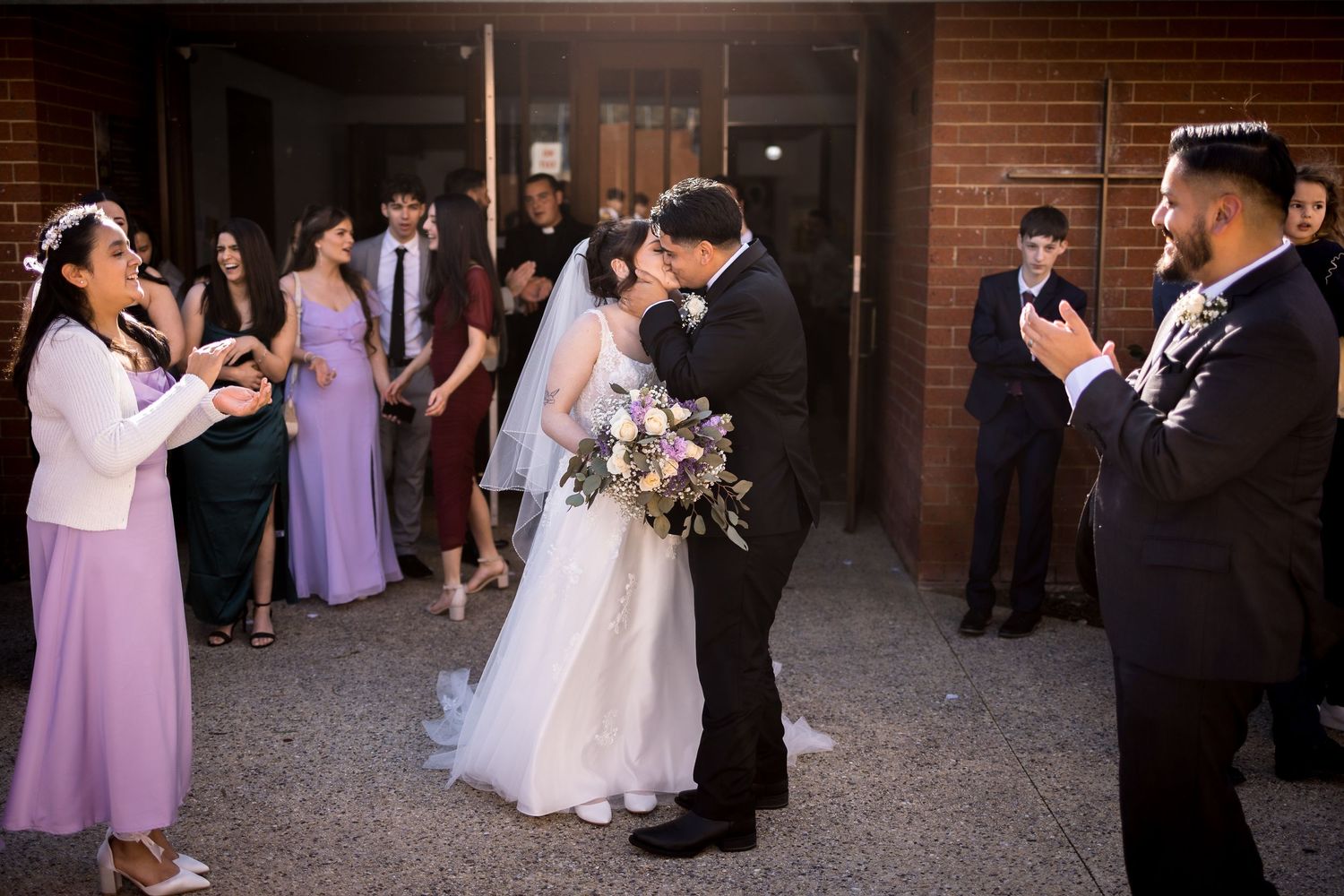 Wedding party gathered outside brick building with bridesmaids in purple dresses.
