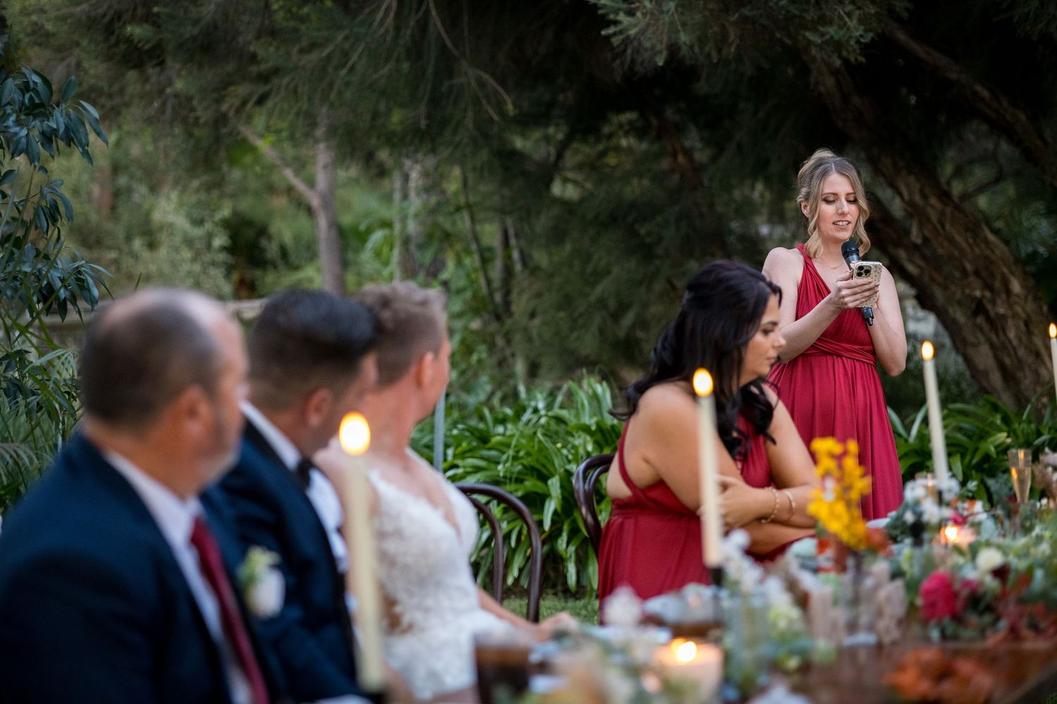 Outdoor wedding reception with guests dining at decorated tables under natural tree canopy.