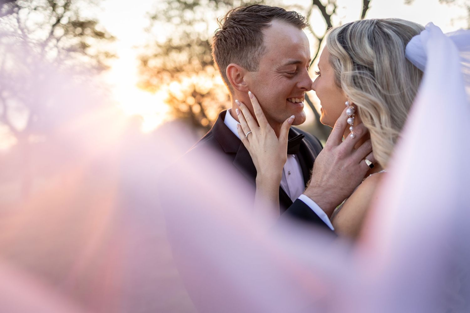 Bride and groom share a romantic sunset moment, framed by soft pink wedding veil, exchanging tender smiles outdoors.
