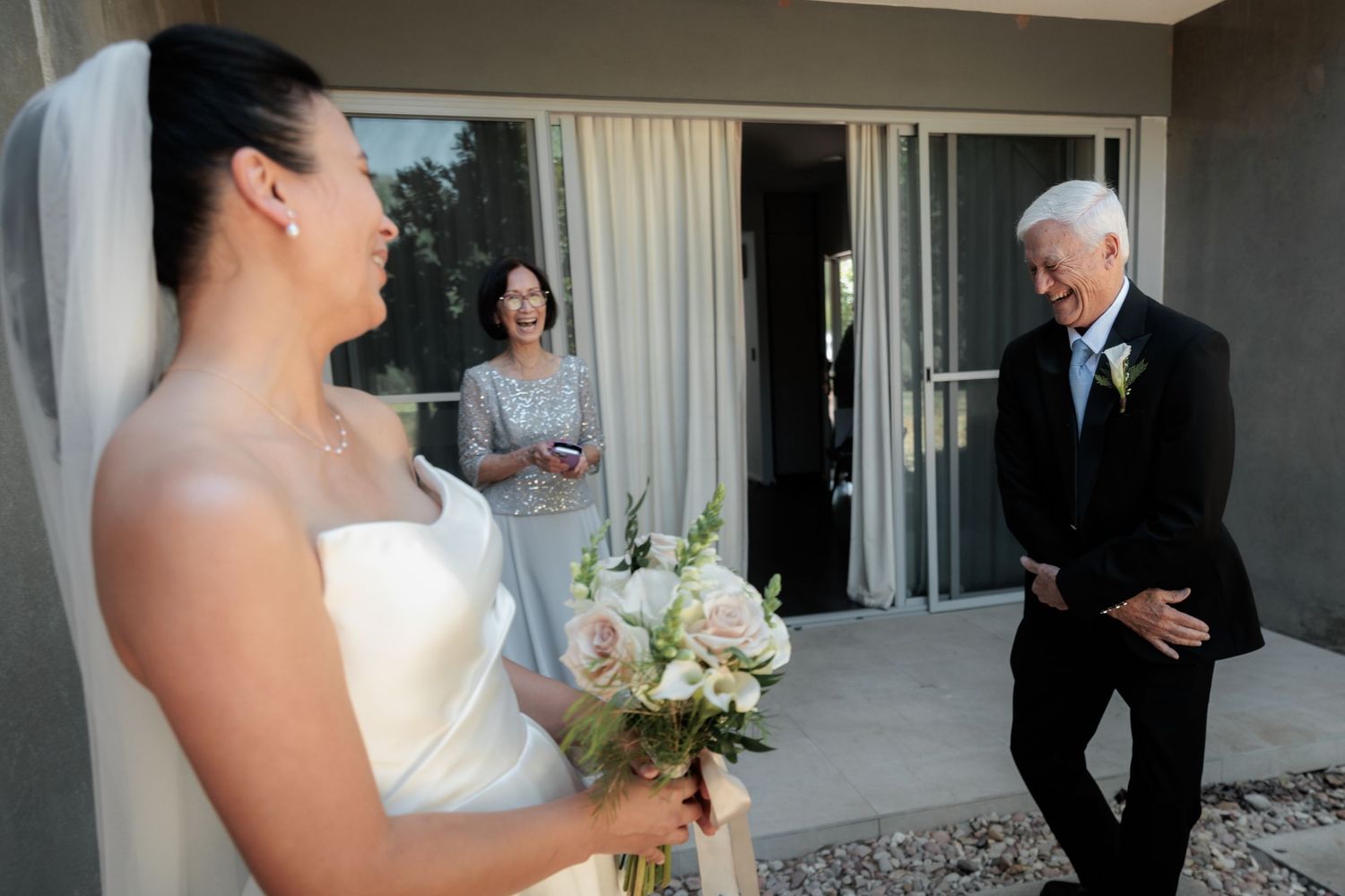 Bride in white gown holds bouquet as emotional father reacts during wedding first look moment outdoors.