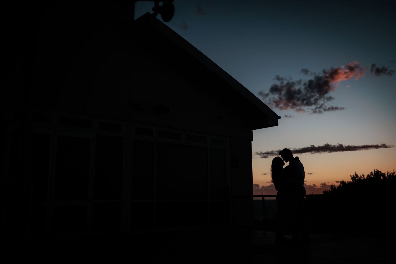 Romantic silhouette of a couple embracing at sunset beside a rustic building with colorful twilight sky.