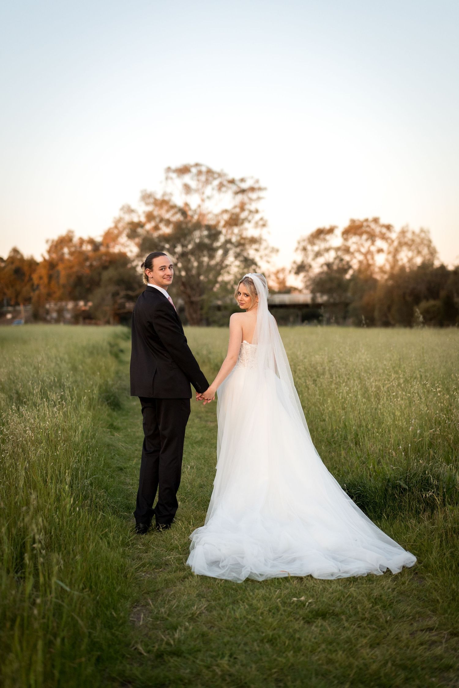 A bride and groom hold hands in a lush green field at sunset, both looking back at the camera in wedding attire.