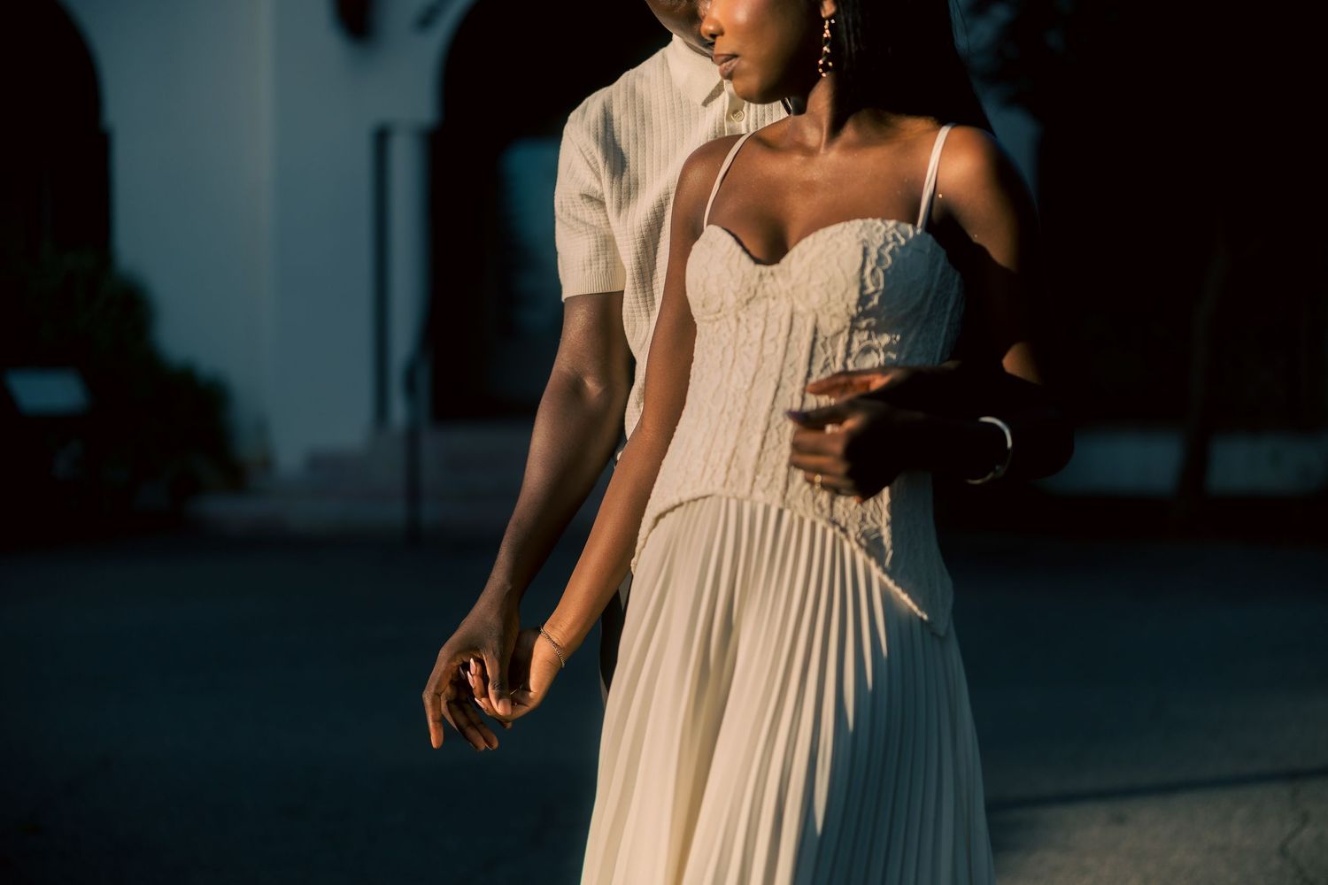 Woman in a pleated cream sundress with spaghetti straps embraced from behind by a person in a light shirt, holding hands in warm sunlight.