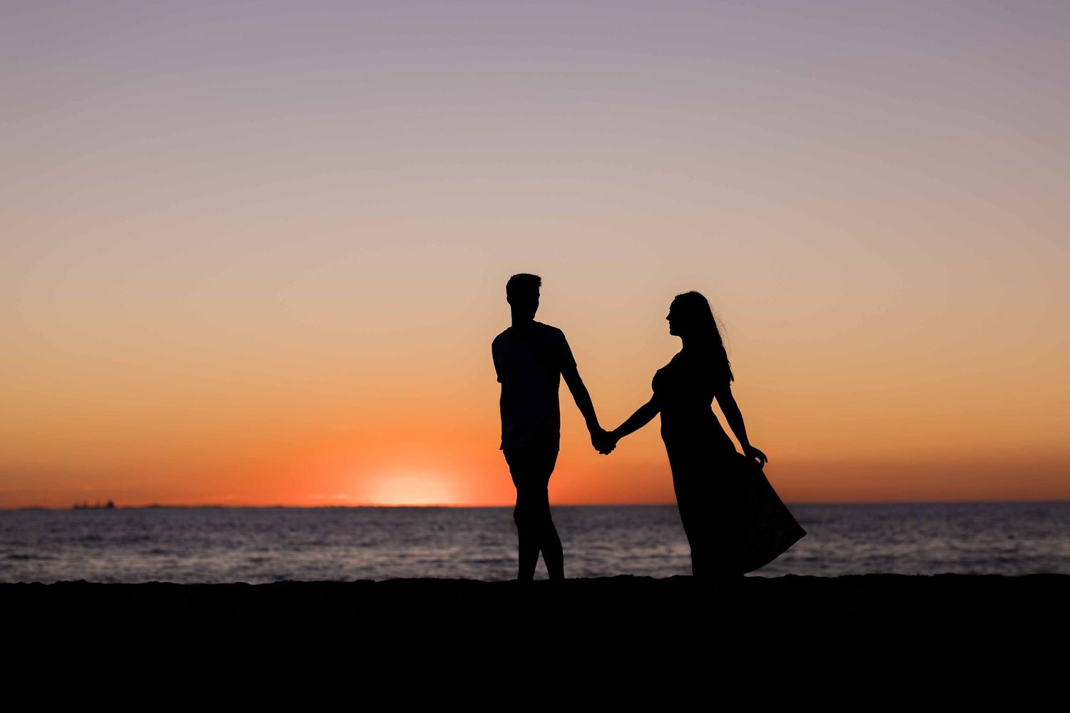 Silhouette of a romantic couple holding hands on a beach at sunset with vibrant orange and purple sky.