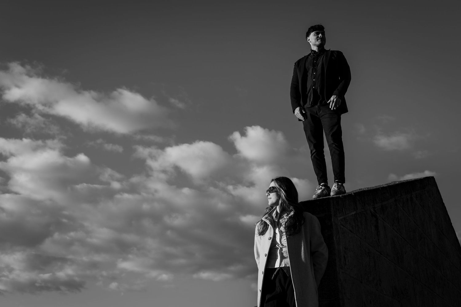 Black and white photo of couple posing dramatically against cloudy sky.