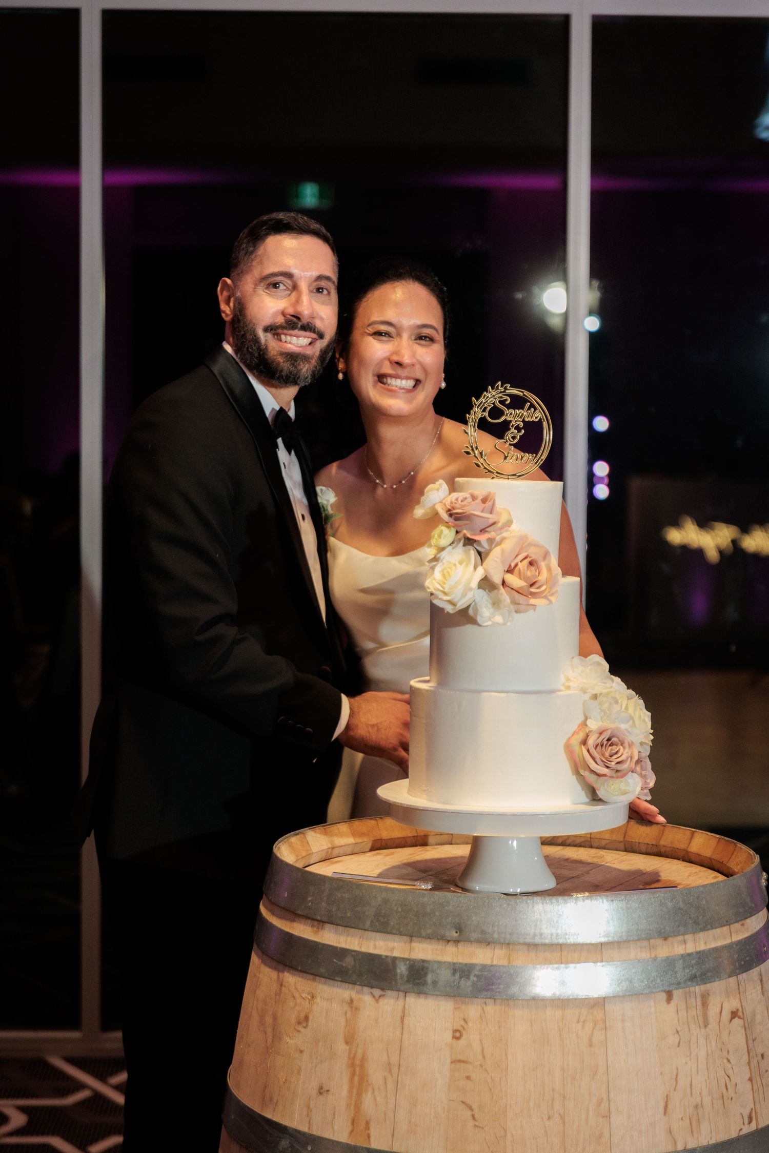 Smiling newlyweds cut their two-tier white wedding cake adorned with roses on a rustic barrel.