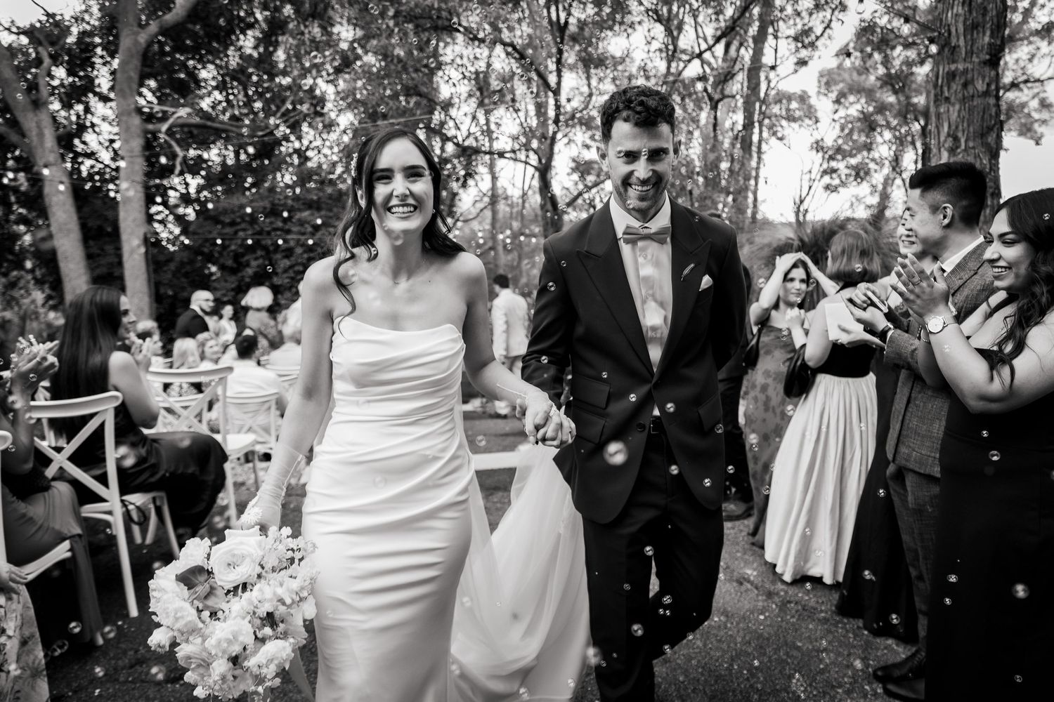 Black and white photo of newlyweds walking back down aisle as guests throw confetti in outdoor ceremony.