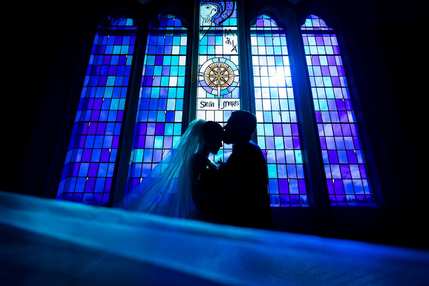 Bride and groom silhouetted in romantic kiss before vibrant blue stained glass church windows.