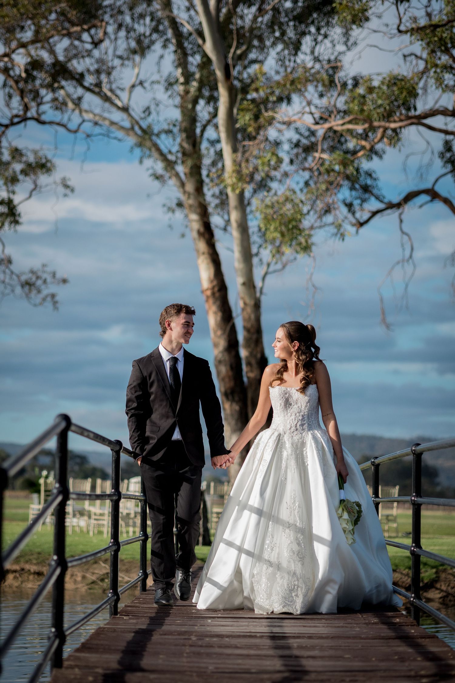 Bride and groom walk hand-in-hand on a wooden bridge, surrounded by lush greenery and a scenic countryside backdrop.