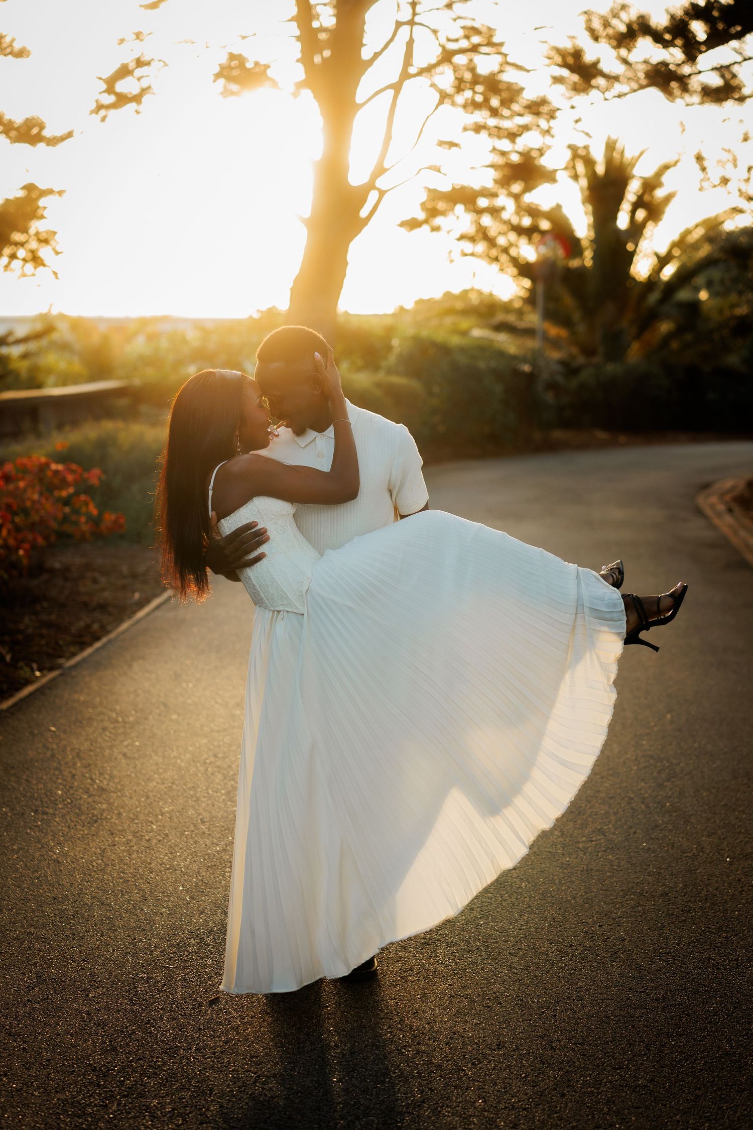 Man in white shirt carries a woman in a flowing white pleated dress along a paved path as the golden sunset glows behind trees.