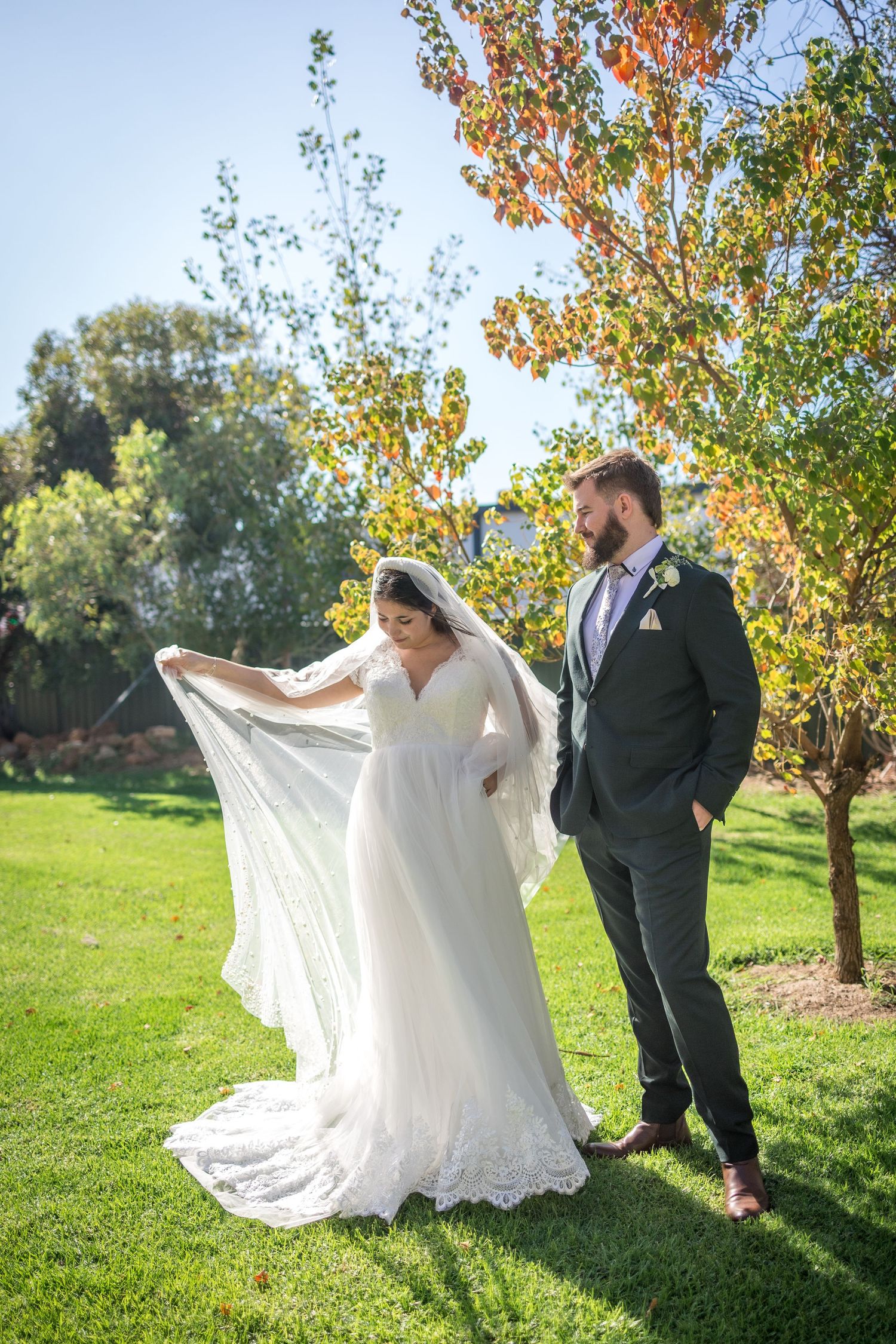 Bride in lace gown lifts veil while groom in dark suit admires her outdoors among autumn trees.