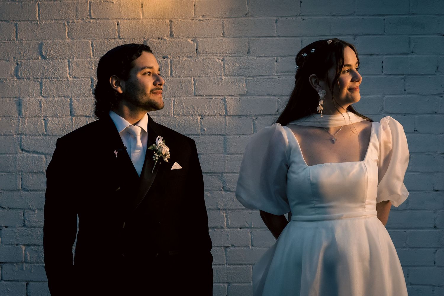 Bride in white gown and groom in black tuxedo posing against a white brick wall in elegant wedding portraits.