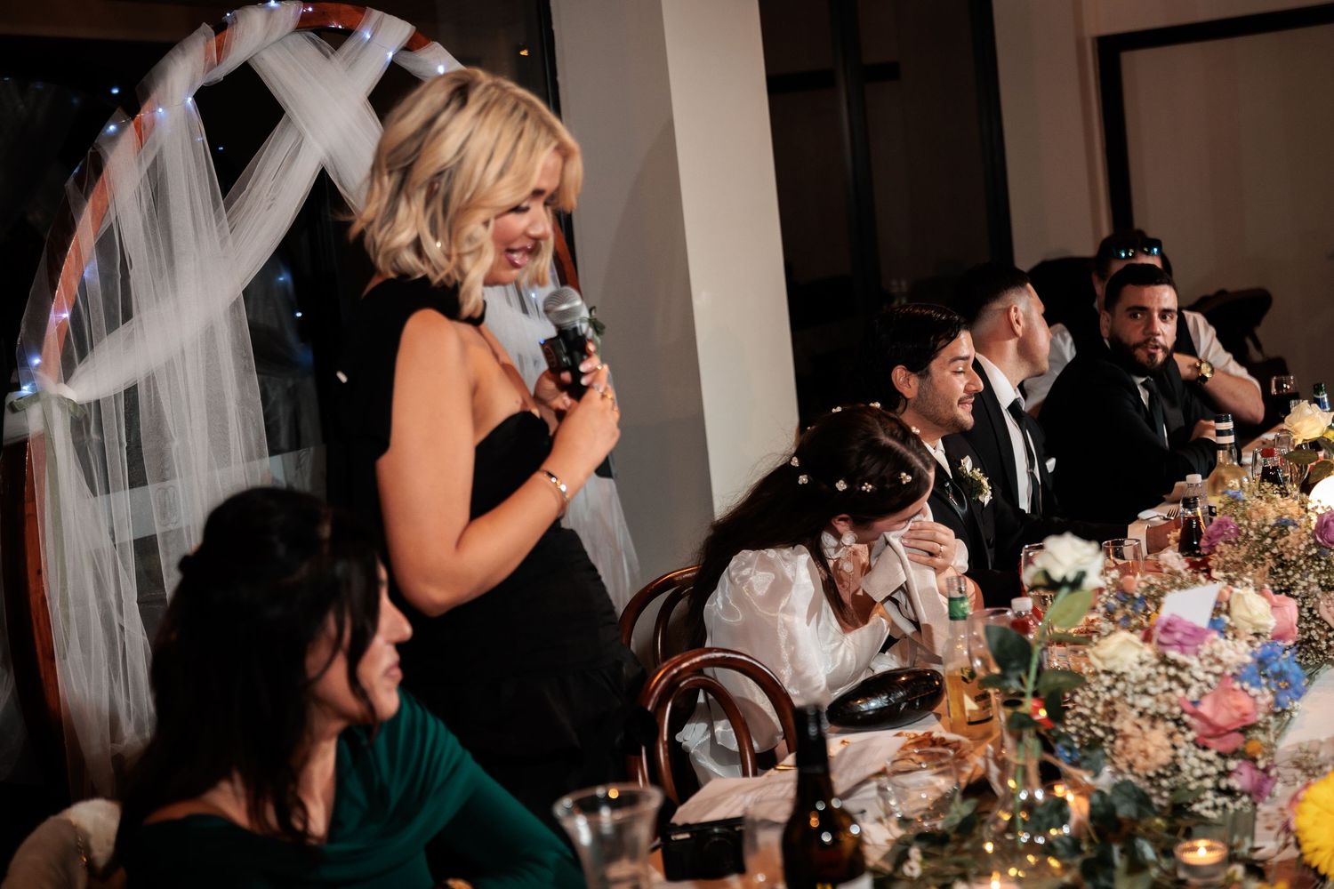 Woman giving a speech with microphone at an elegant wedding reception dinner with guests seated at a floral decorated table.