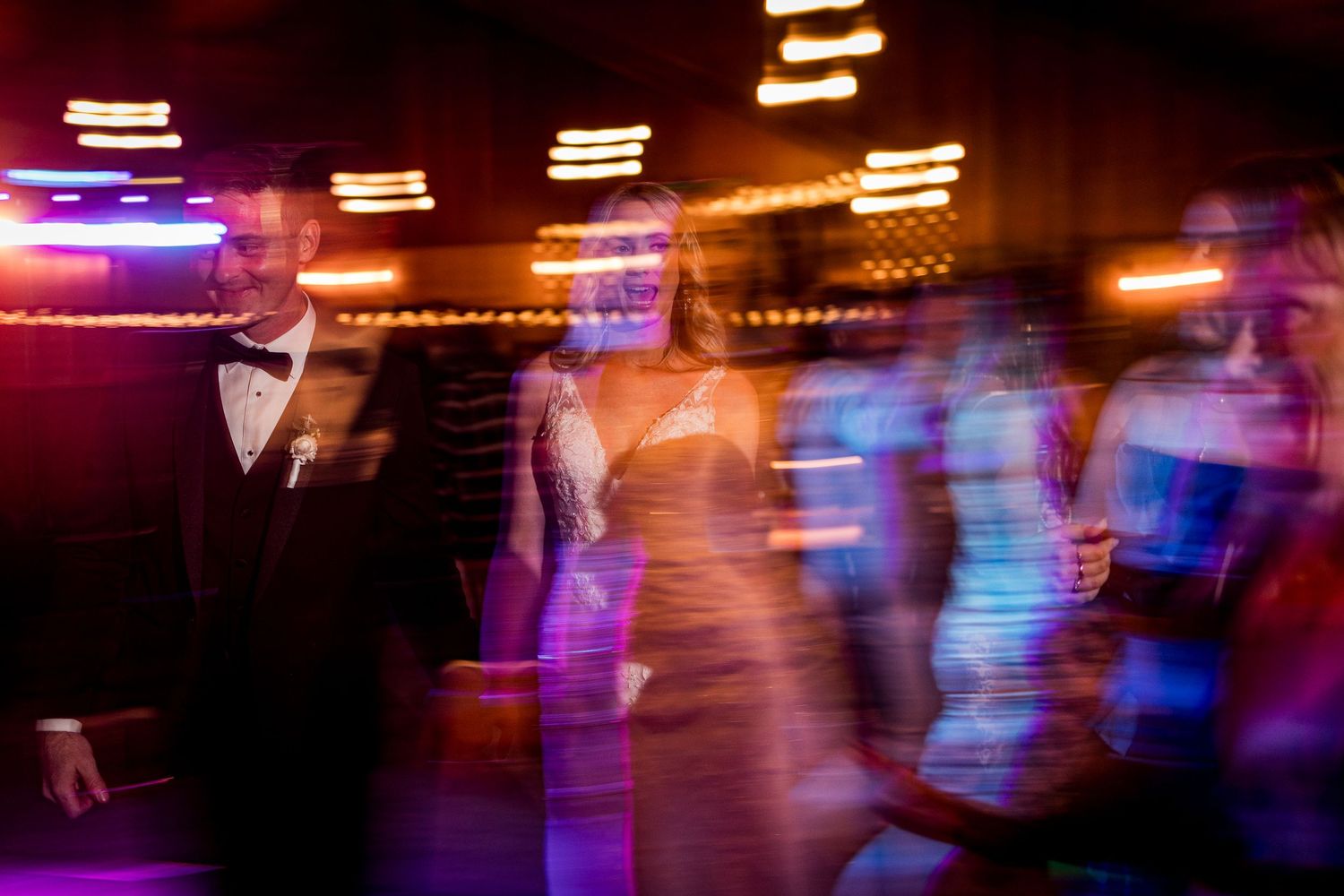 Blurred motion shot of elegantly dressed wedding guests dancing under colorful lights at a festive reception.
