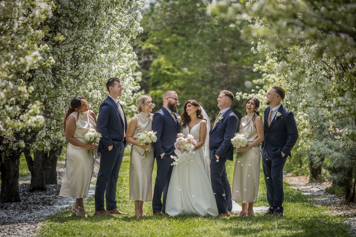 Wedding party poses outdoors among blooming white trees, bride in white gown with bouquet, bridesmaids in champagne dresses.
