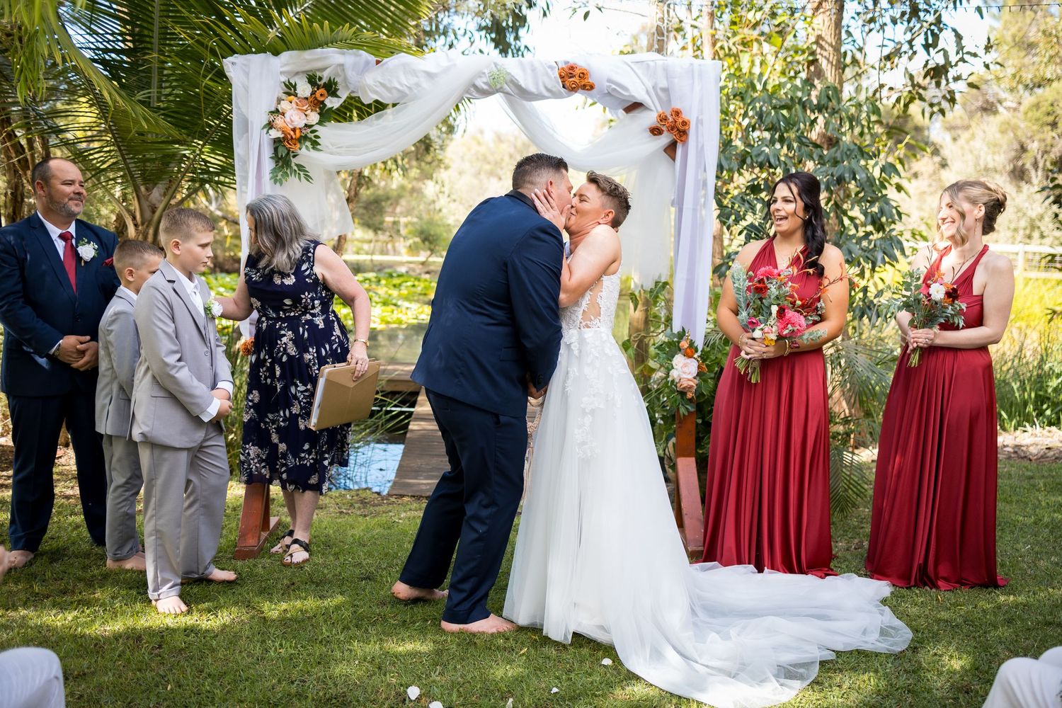 Garden wedding ceremony with bridesmaids in red dresses and floral arch decorations.
